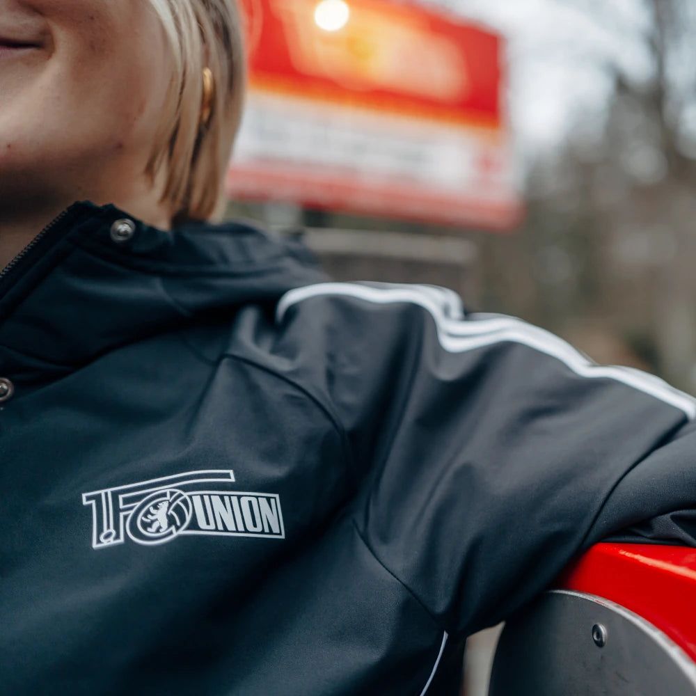 Eine Person in einer sportlichen Jacke mit dem Logo "1.FC UNION" lehnt an einer Sitzgelegenheit. Hintergrund unscharf.