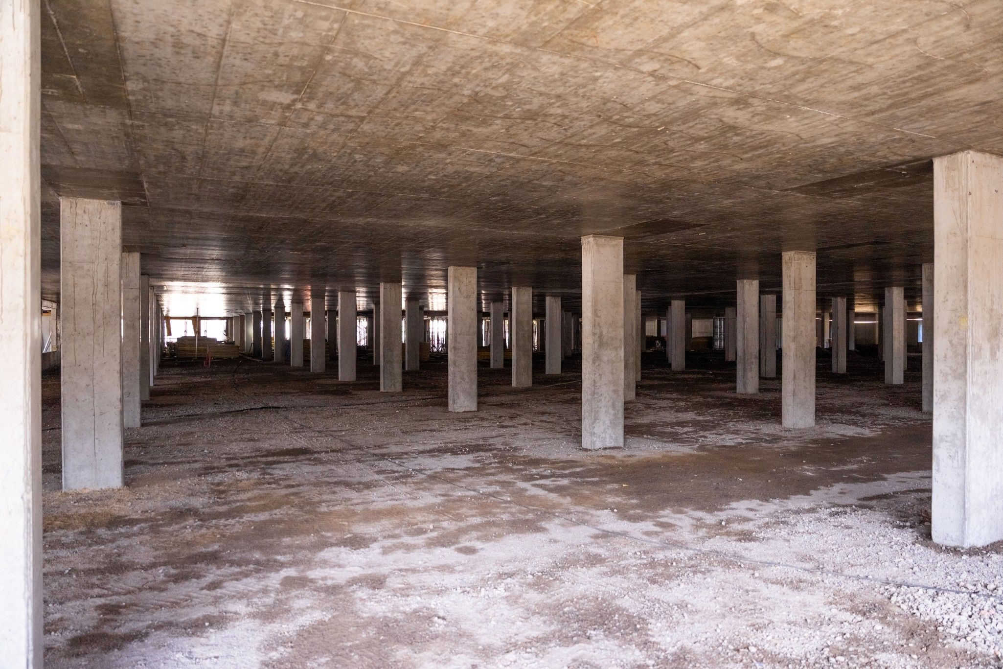 Innenansicht eines Rohbaus mit Sichtbetonsäulen und einem unvollendeten Boden, der mit Schutt und Staub bedeckt ist.