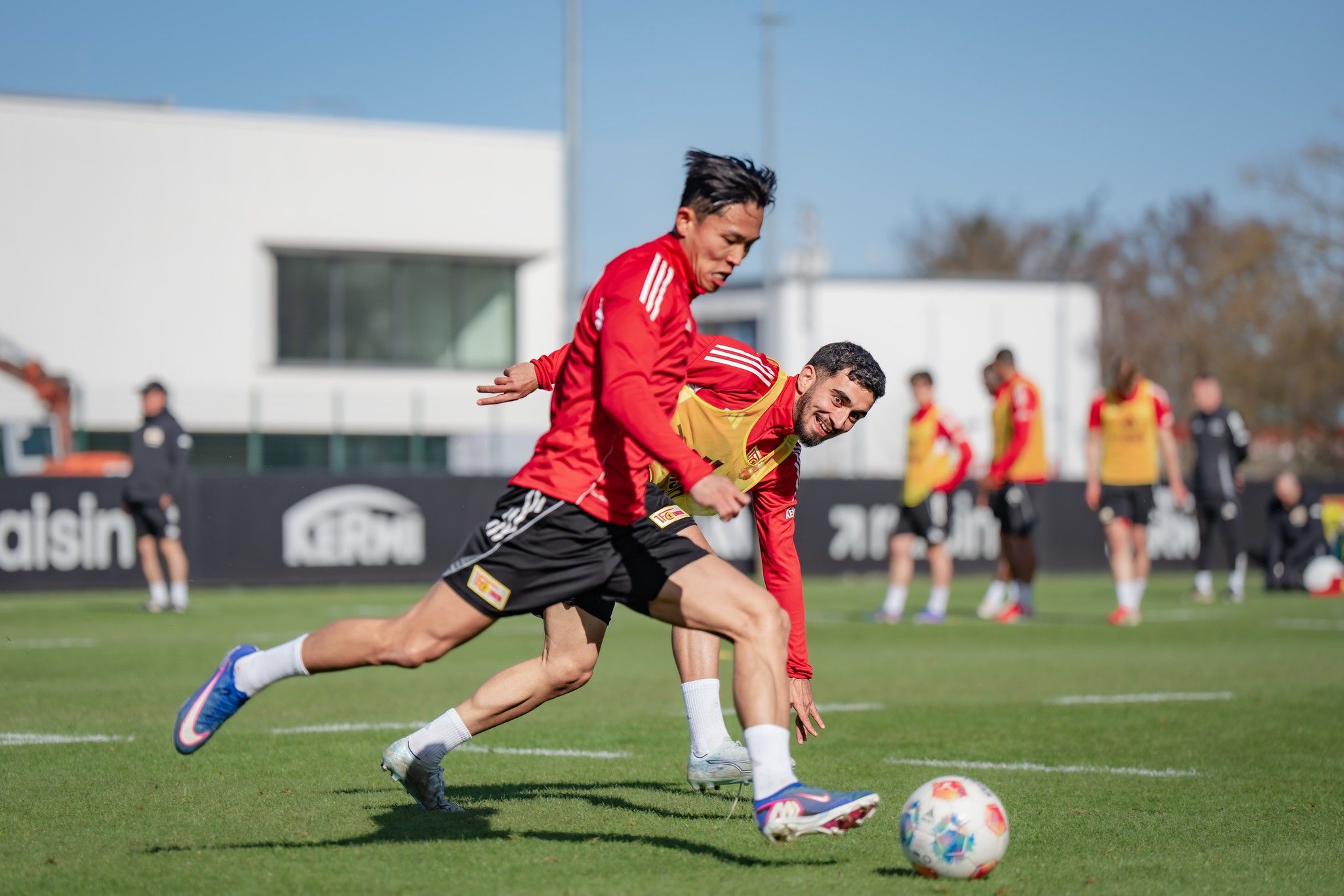 Zwei Fußballspieler in roten Trikots trainieren auf einem grünen Kunstrasenplatz, während andere Spieler im Hintergrund Übungen durchführen.