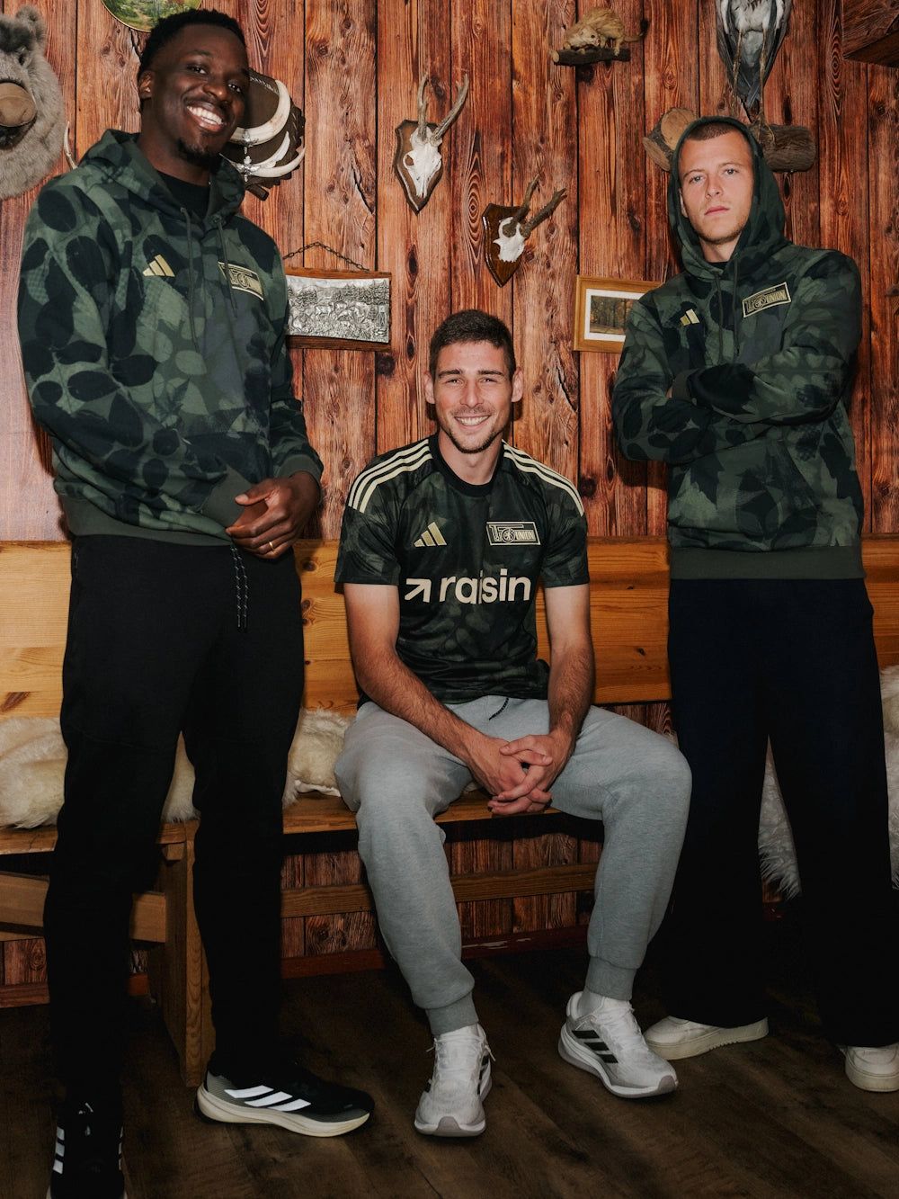 Drei Männer in sportlicher Kleidung stehen und sitzen vor einer Holzwand mit Tierpräparaten.