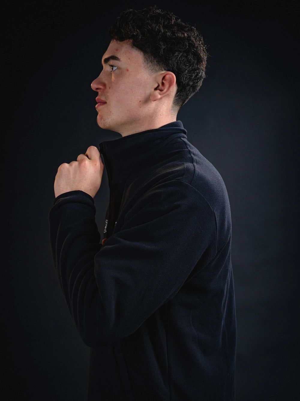 Ein junger Mann in einem dunklen Fleece-Oberteil, der nachdenklich zur Seite schaut und seinen Kragen hochzieht.