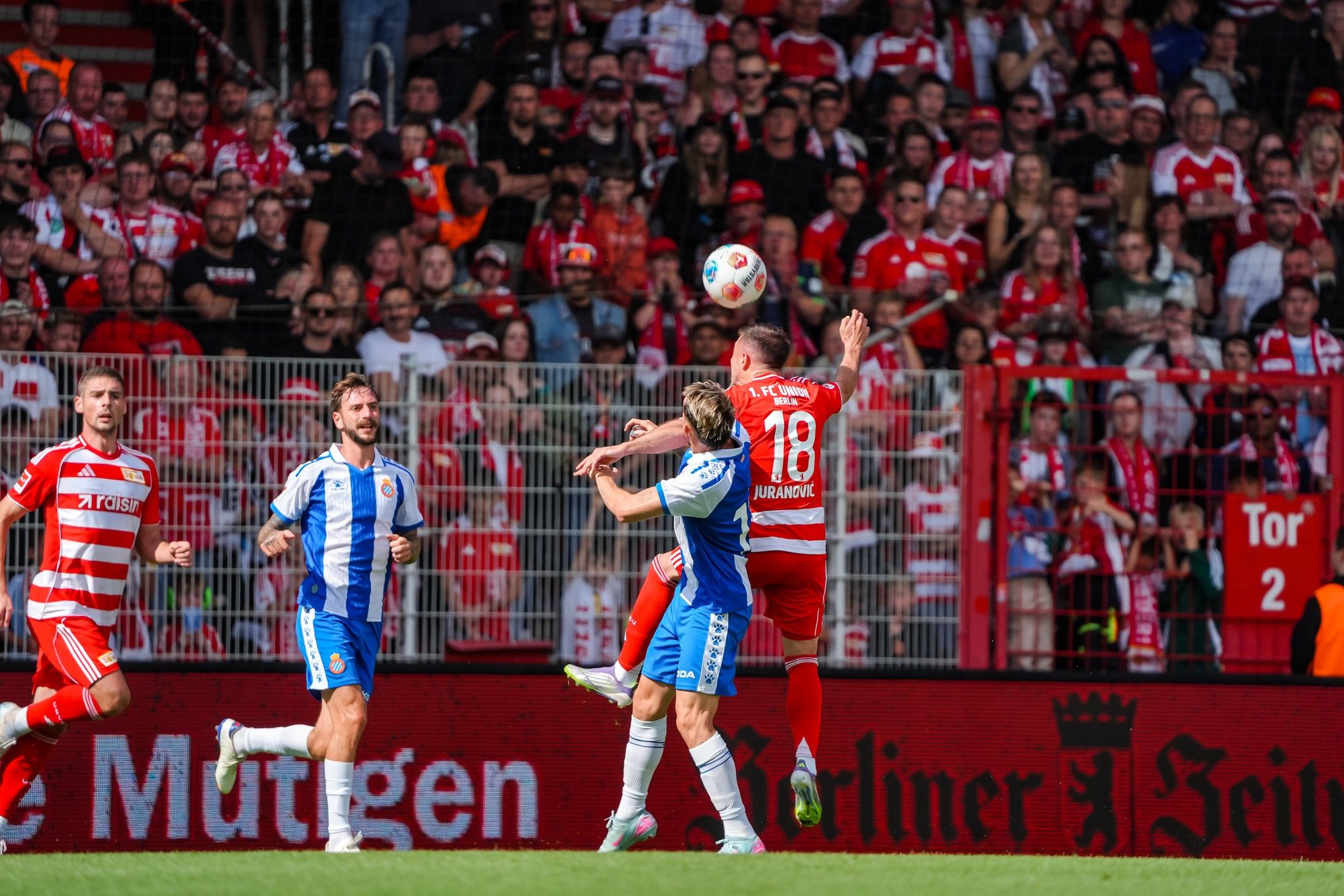 Ein Fußballspiel mit Spielern in roten und blauen Trikots, während ein Spieler den Ball mit dem Kopf spielt und die Fans jubeln.
