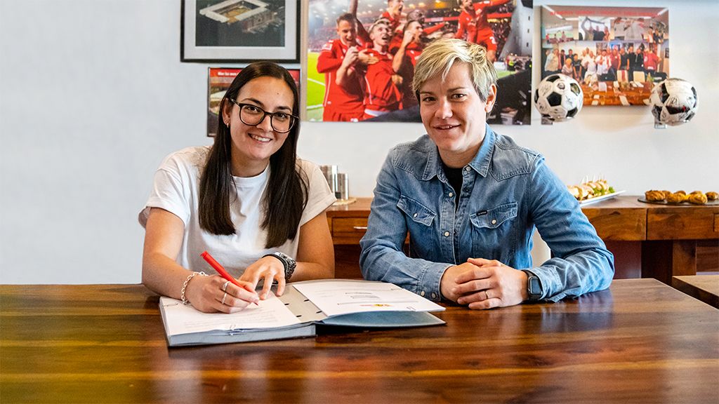 Eine junge Frau unterschreibt einen Vertrag am Tisch, während eine andere Frau lächelnd daneben sitzt. Im Hintergrund sind Fußballbilder und -utensilien.
