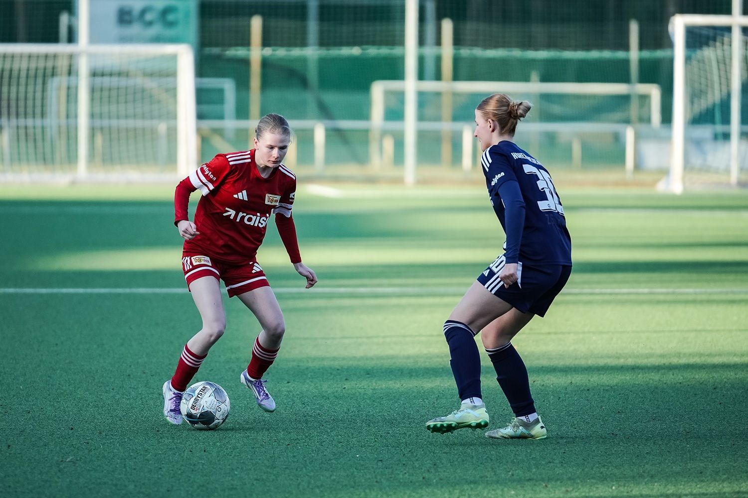 Zwei Fußballspielerinnen auf einem Kunstrasenplatz im Wettkampf um den Ball.