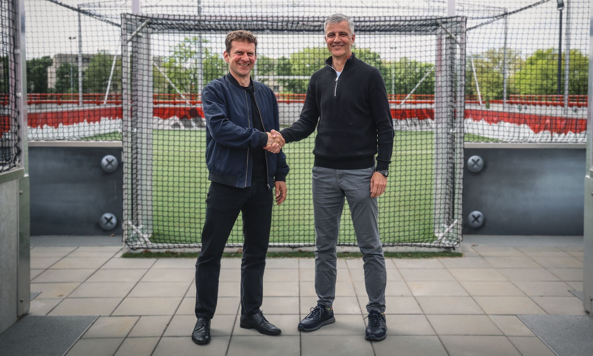 Zwei Männer stehen auf einem Sportfeld und schütteln sich die Hände, im Hintergrund ein Fußballtor.