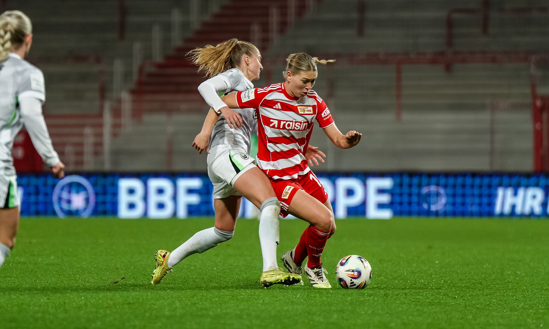 Spielszene im Frauenfußball: Eine Spielerin in rot-gestreiftem Trikot dribbelt den Ball, während eine Gegenspielerin in Grau angreift.