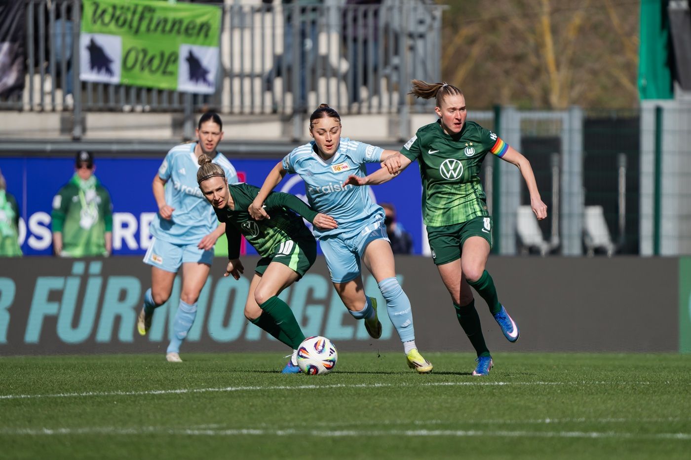 Zwei Fußballspielerinnen kämpfen um den Ball, während weitere Spielerinnen im Hintergrund agieren. Spielszene auf dem Rasen.