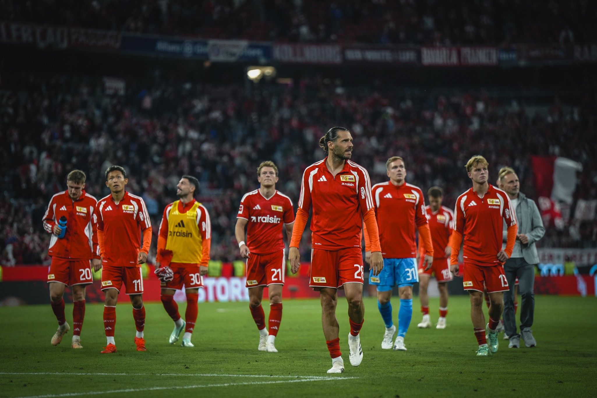 Eine Gruppe von Fußballspielern in roten Trikots geht nach einem Spiel über das Spielfeld, im Hintergrund jubelnde Fans.