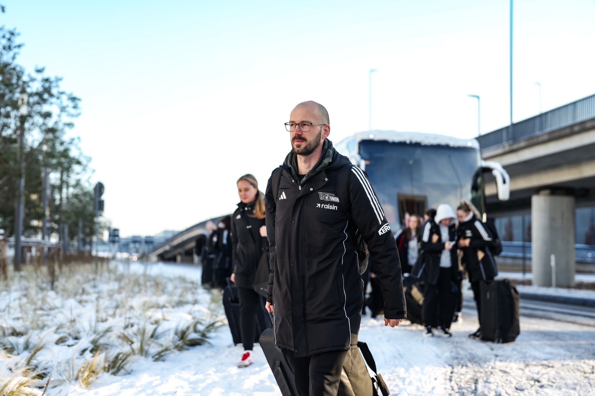 Gruppe von Personen, die mit Gepäck auf einem verschneiten Weg in Richtung eines Busses gehen.