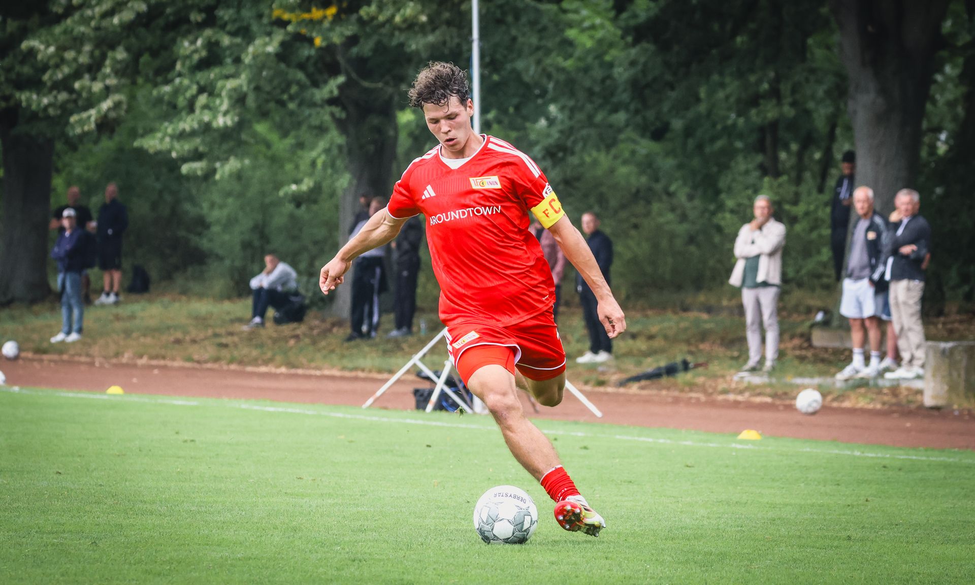 Ein Fußballspieler in rotem Trikot führt einen Schuss auf dem Platz aus, während Zuschauer im Hintergrund stehen.