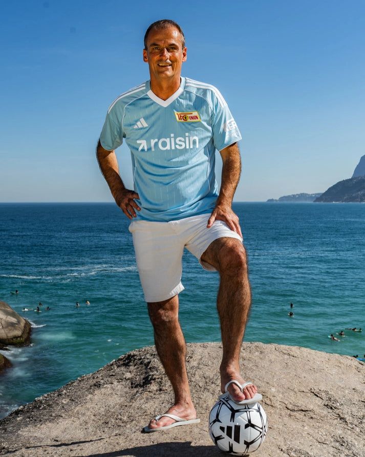 Mann in einem hellblauen Fußballtrikot steht barfuß auf einem Felsen mit einem Fußball, der Ozean im Hintergrund.