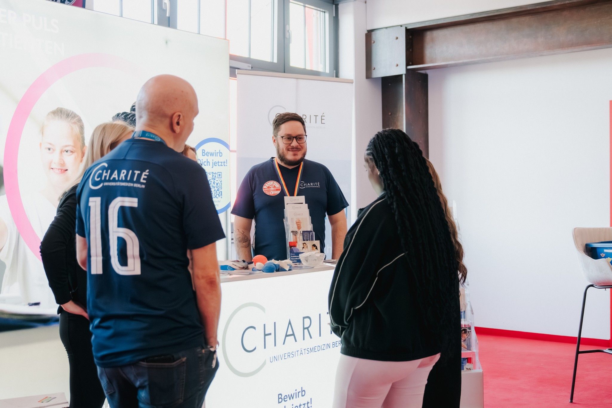 Personen stehen an einem Informationsstand der Charité, während ein Mitarbeiter sie berät und Informationsmaterial verteilt.
