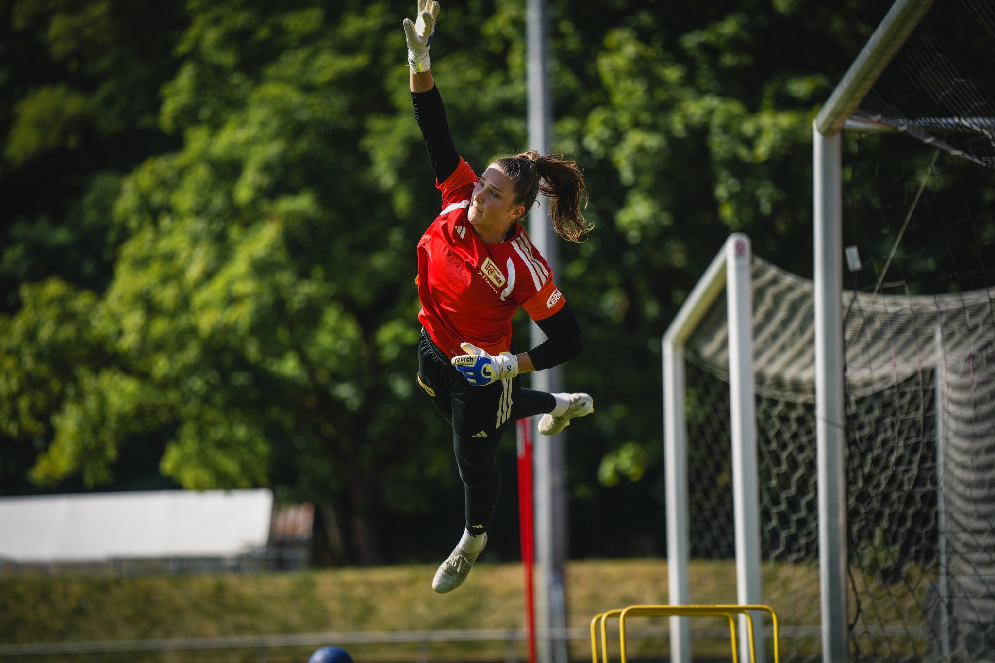 Eine Fußballtorhüterin springt in einem roten Trikot, um einen hohen Ball zu parieren, vor einem Tor auf einem Sportplatz.