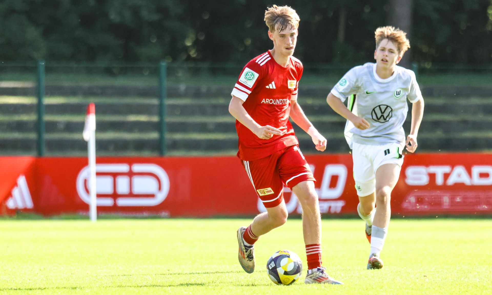 Zwei Jugendliche spielen Fußball auf einem Rasenplatz, der Spieler in Rot dribbelt den Ball, während der in Weiß ihn verfolgt.