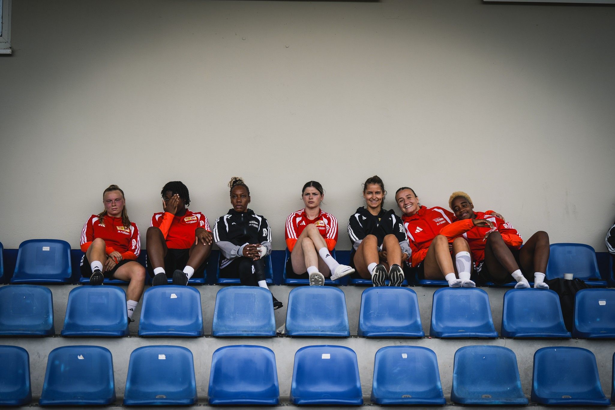 Eine Gruppe junger Sportlerinnen in roten Trainingsanzügen sitzt entspannt auf blauen Sitzen in einem Stadion.