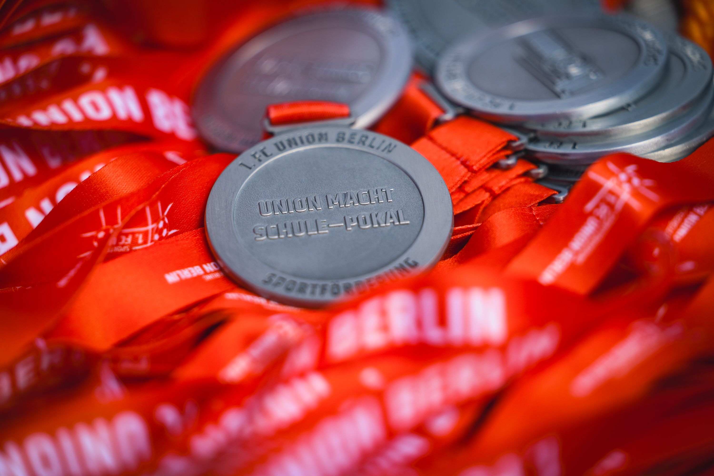 Medaille mit orangem Band, bedruckt mit "UNION MACHT SCHULE-POKAL", umgeben von weiteren Medaillen in einer Box.