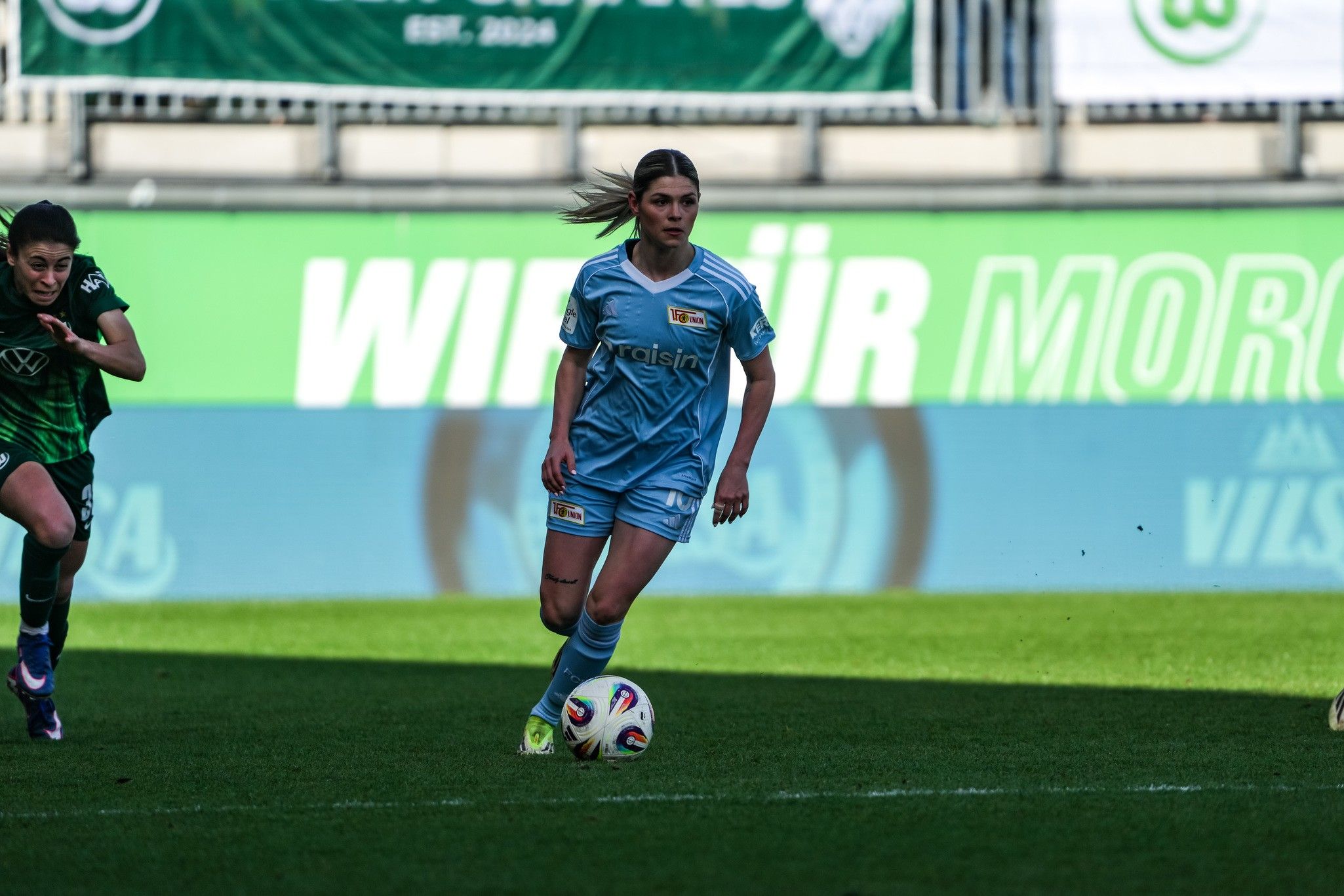 Eine Spielerin in blauer Fußballausrüstung dribbelt den Ball auf einem Fußballfeld, während eine Gegnerin in grüner Ausrüstung nahe ist.