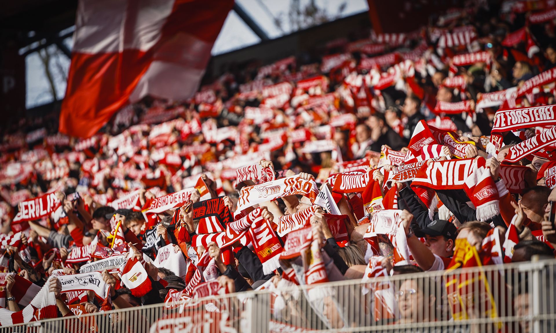 Eine große Menge von Fans in einem Stadion, die rote Schals und Fahnen schwenken und eine enthusiastische Stimmung erzeugen.