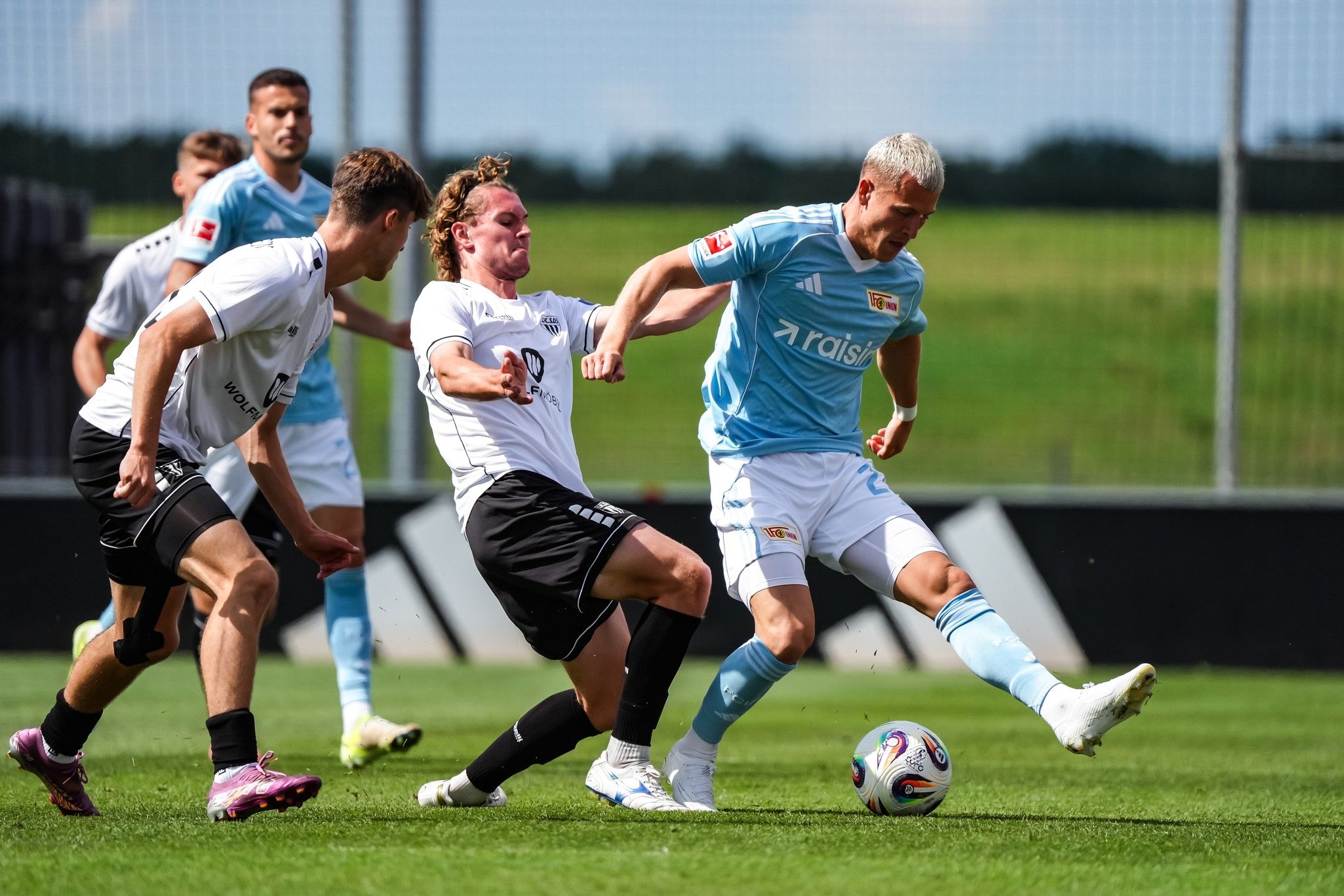Zwei Fußballspieler kämpfen um den Ball, während ein dritter Spieler in der Nähe steht. Helle, sonnige Atmosphäre auf dem Spielfeld.