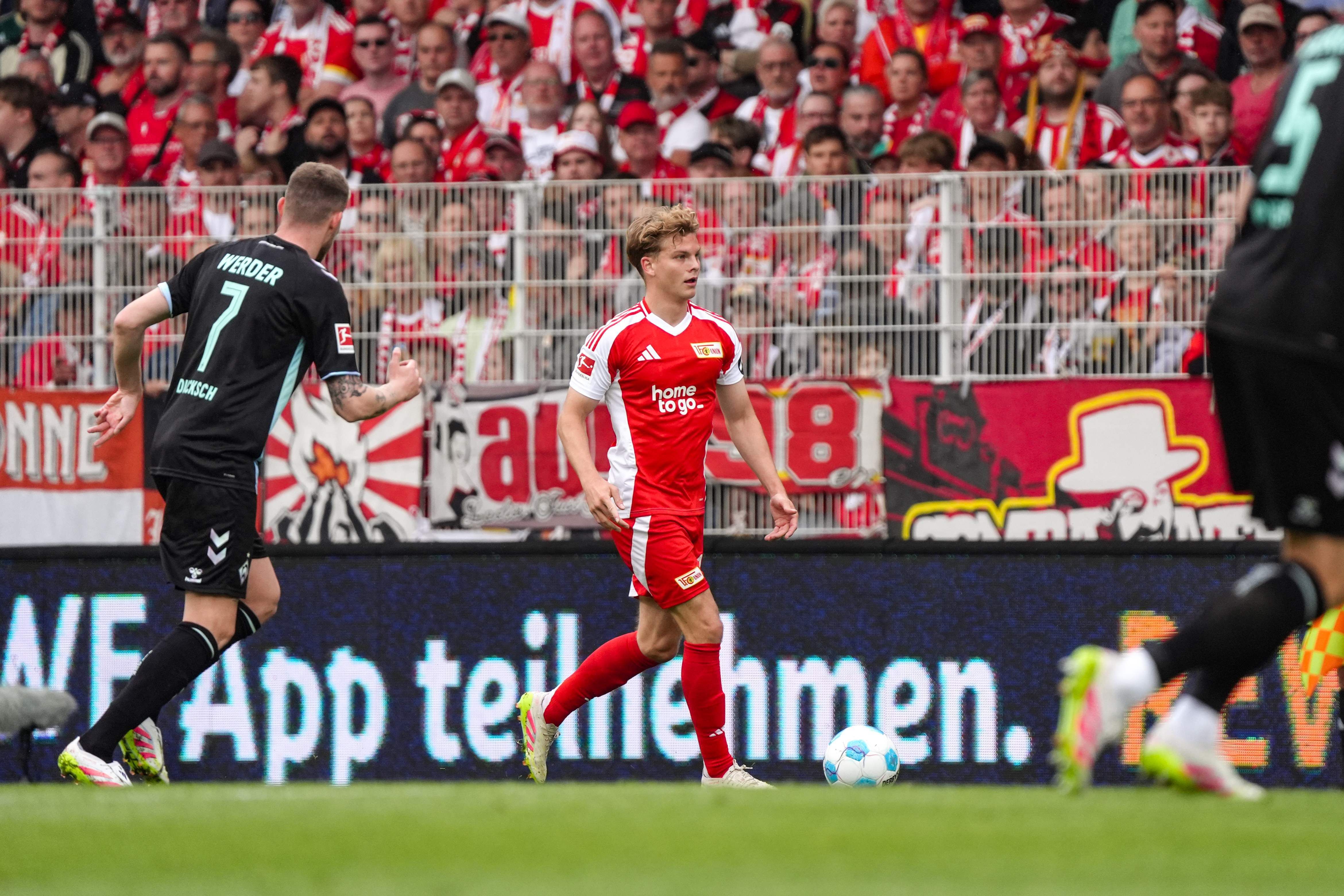 Ein Fußballspiel mit Spielern in roten und schwarzen Trikots. Im Hintergrund Fans mit Flaggen und Bannern.
