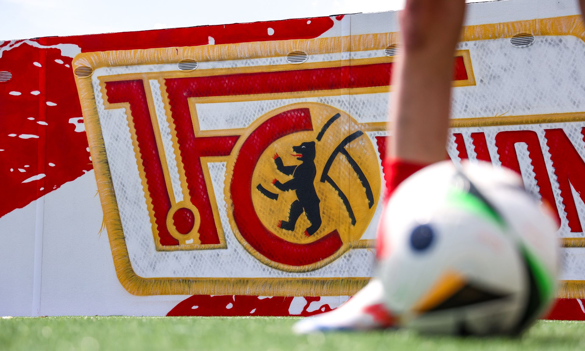 Ein Fuß im Vordergrund neben einem Fußball, vor einem großen Logo des 1. FC Union Berlin mit einem Bären.