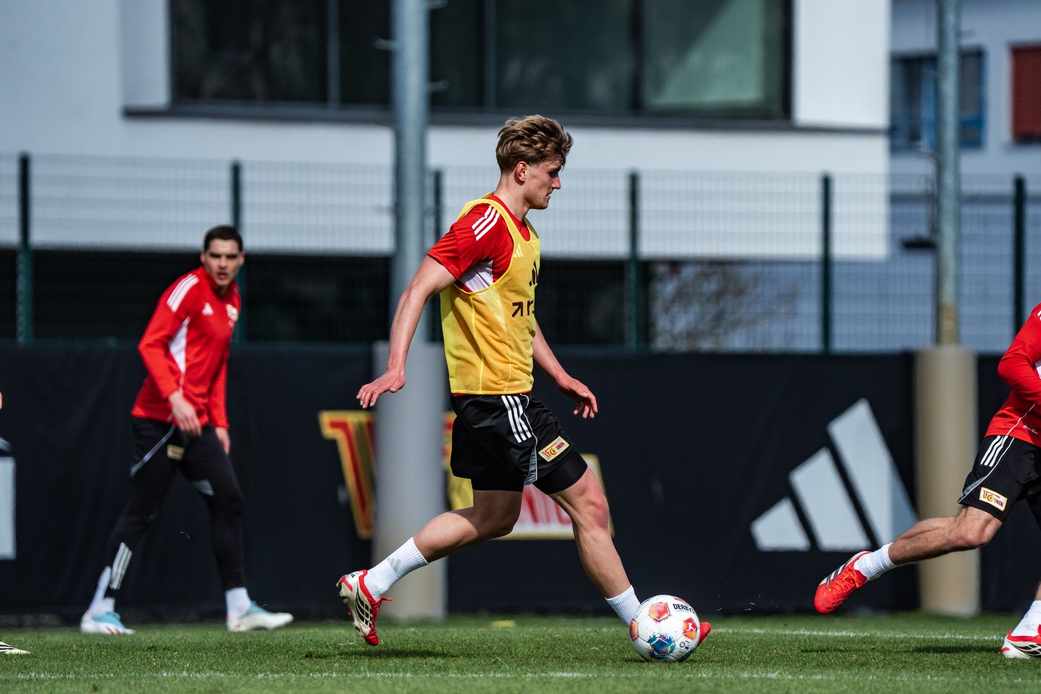 Ein Fußballspieler in gelber Weste dribbelt den Ball auf einem Trainingsplatz, während andere Spieler im Hintergrund aktiv sind.