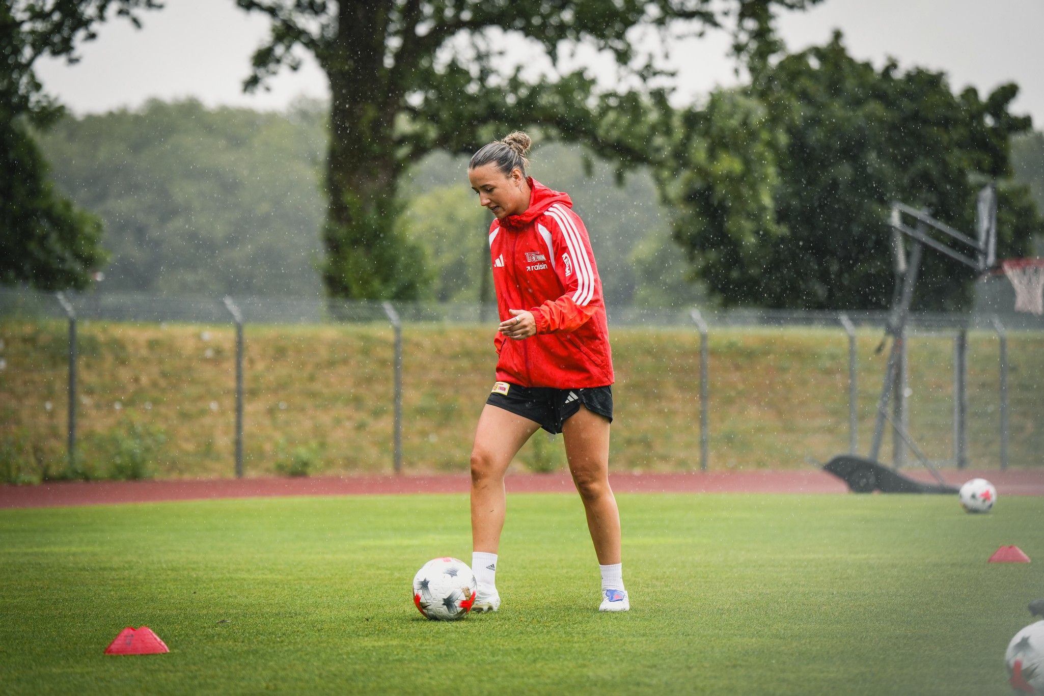 Eine Fußballspielerin in roter Trainingsjacke übt im Regen auf einem Rasenplatz mit einem Fußball.