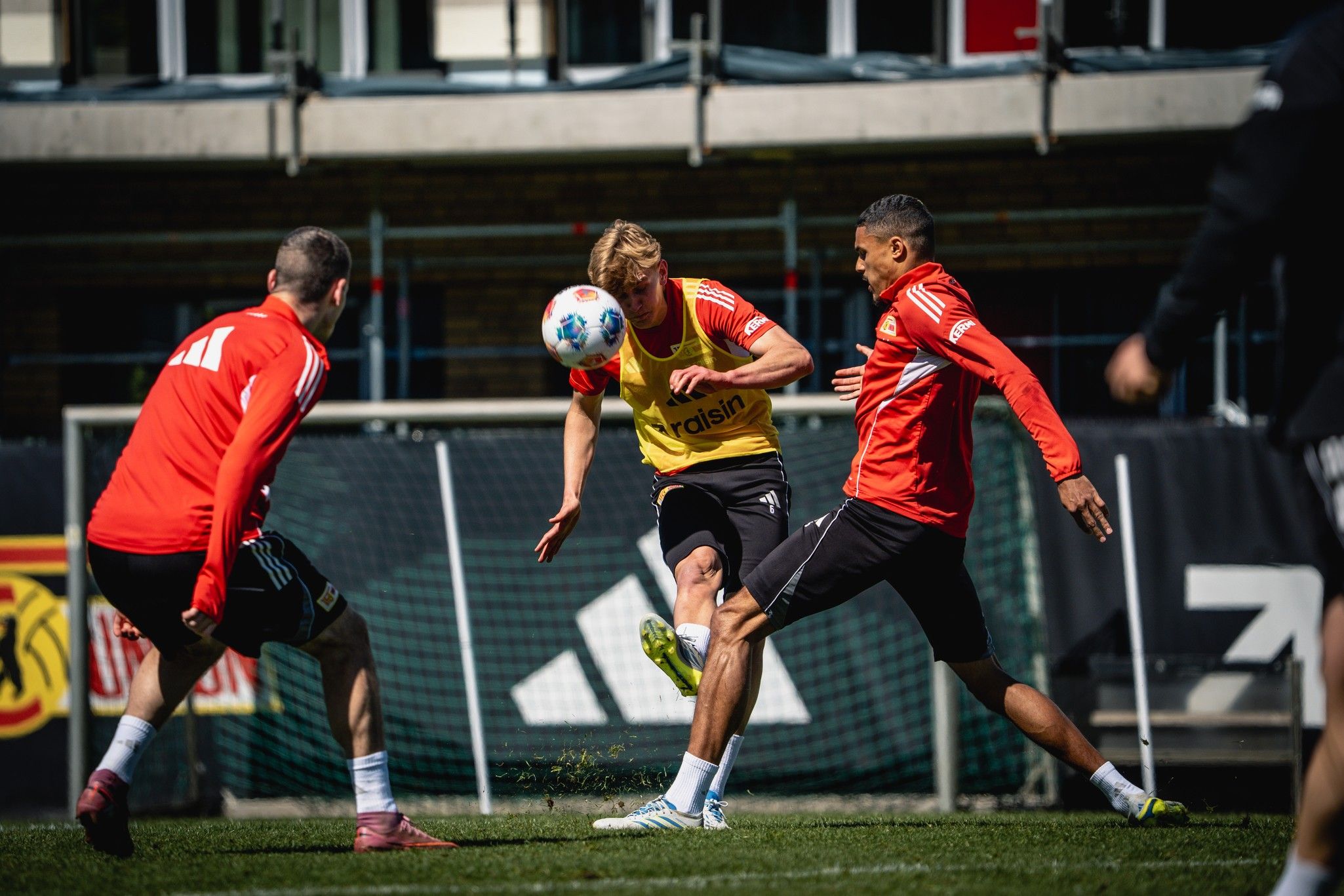 Drei Fußballspieler im Training, einer tritt den Ball, die anderen zwei reagieren auf die Szene. Helle, sportliche Umgebung.