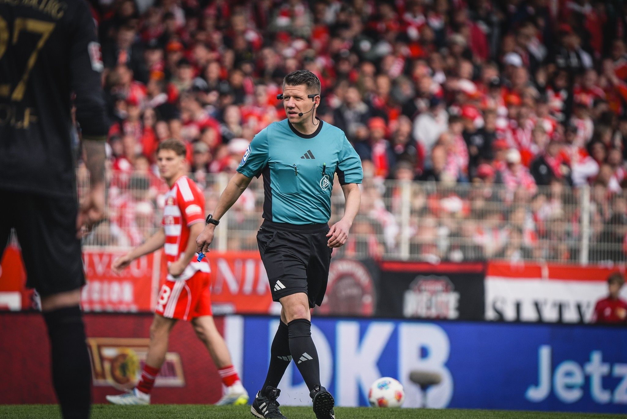 Ein Schiedsrichter in blauem Trikot auf dem Fußballfeld, während Spieler im Hintergrund agieren und Zuschauer im Stadion zu sehen sind.