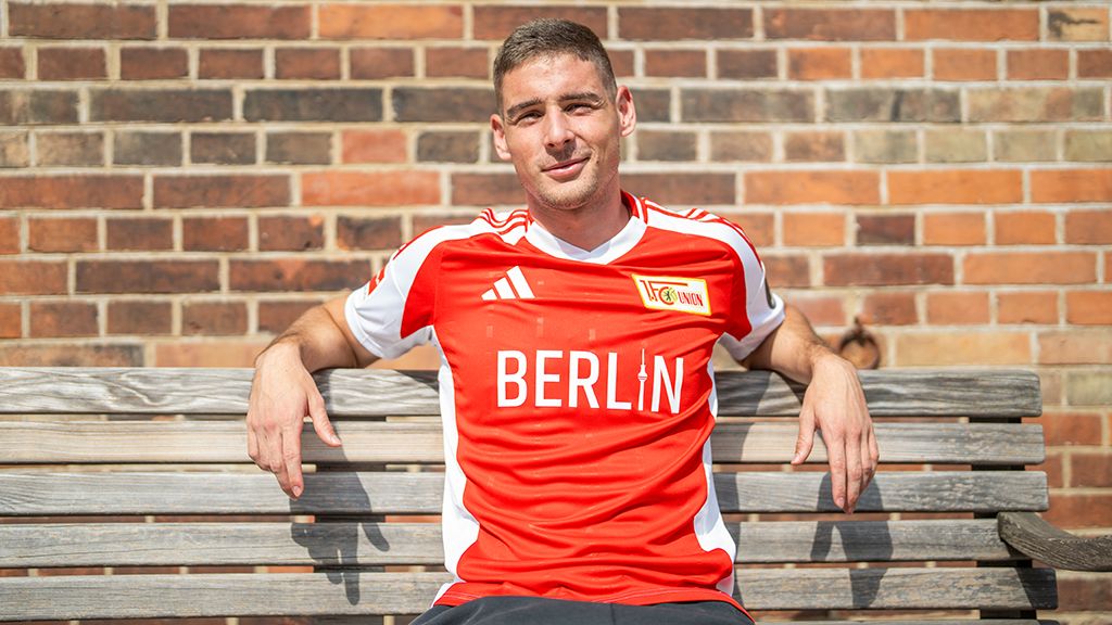 A football player in a red jersey with "BERLIN" written on it is sitting relaxed on a wooden bench in front of a brick wall.