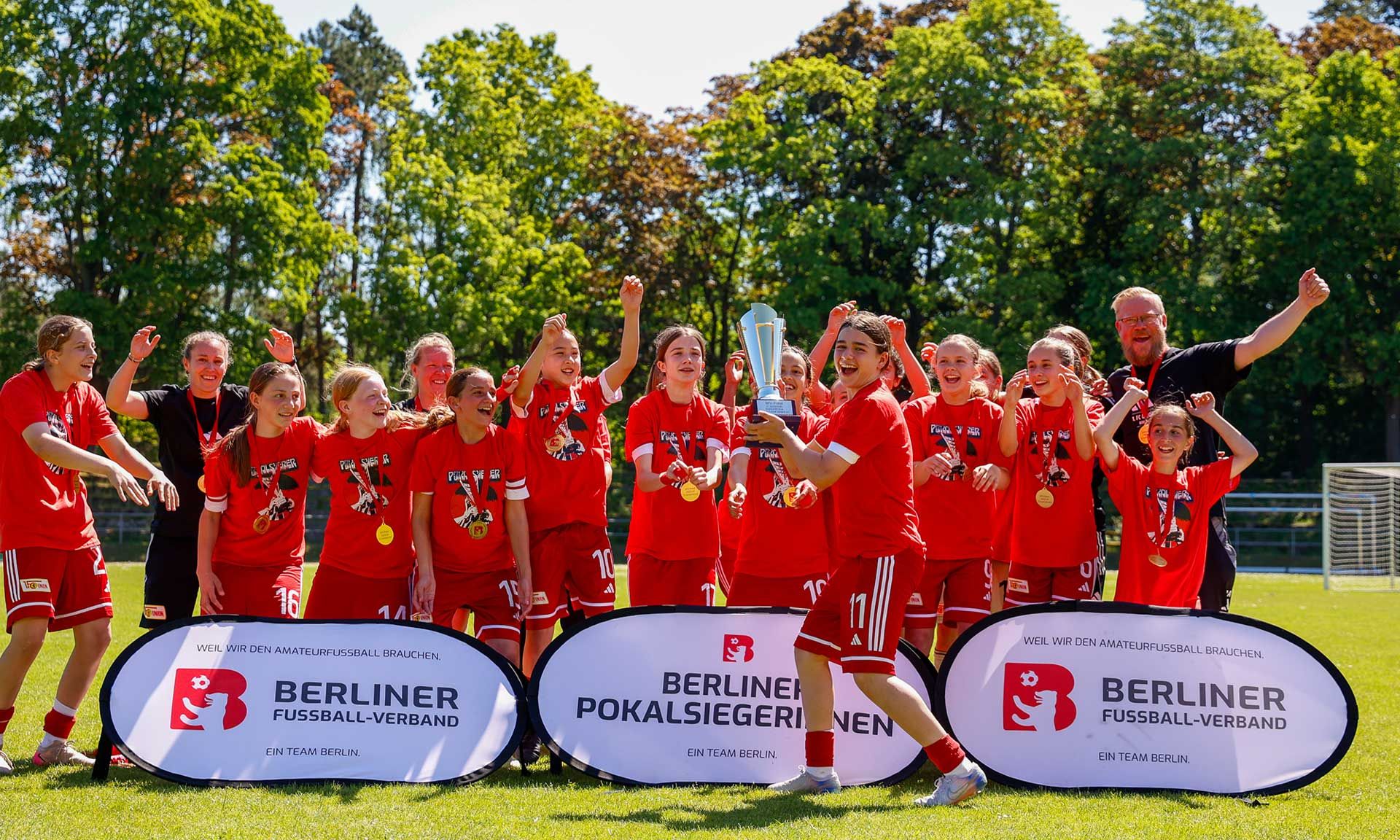 Mädchenfußballmannschaft feiert den Sieg mit einem Pokal, umgeben von Medaillen und Banner des Berliner Fußballverbands.
