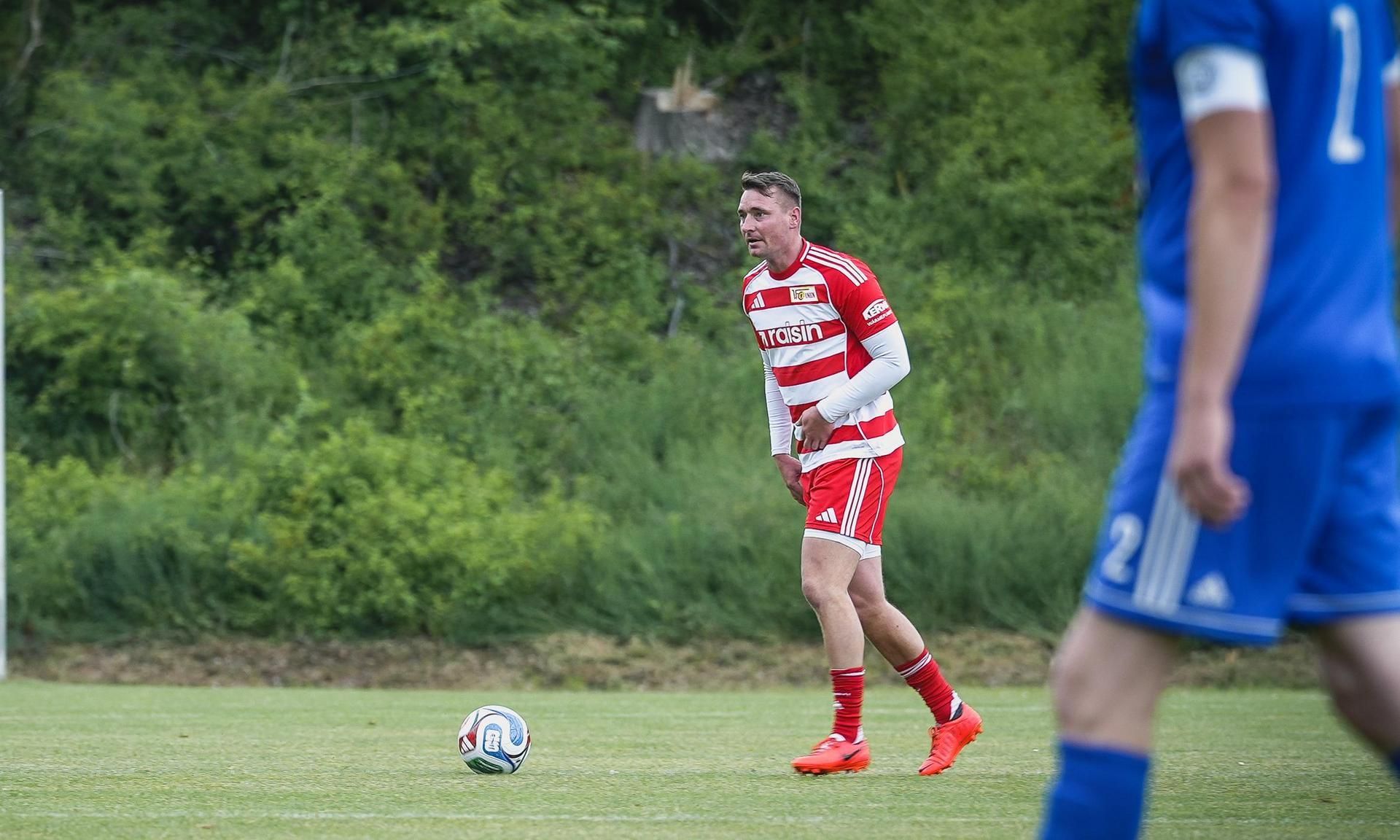 Spieler in rot-weißem Trikot steht auf dem Fußballfeld, bereit für einen Freistoß, während ein gegnerischer Spieler in Blau vorbeiläuft.