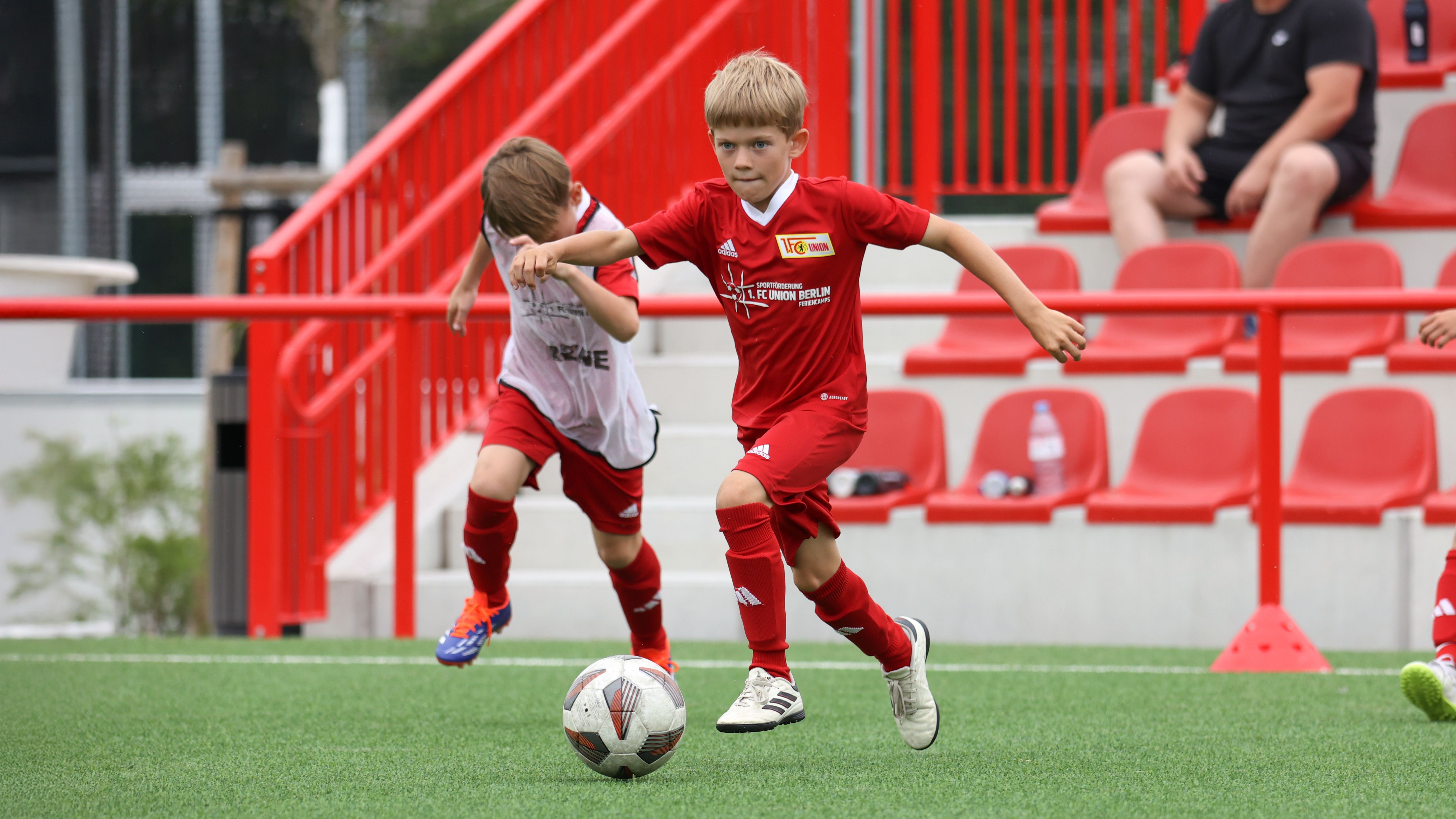 Zwei Kinder in roten Fußballtrikots spielen auf einem Sportplatz; eines dribbelt mit dem Ball, während das andere hinterherläuft.