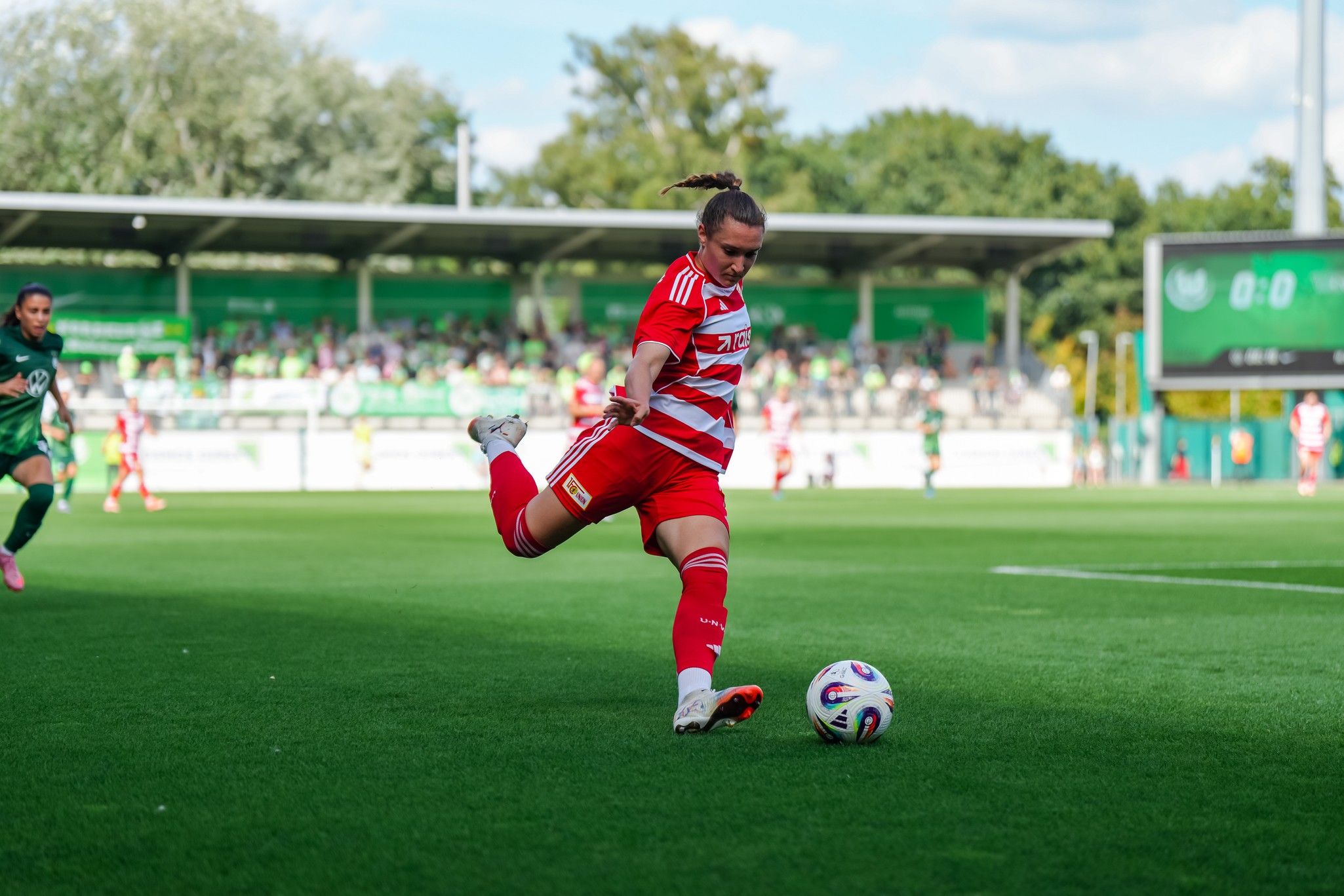 Spielerin in rotem Trikot schießt den Ball auf einem Fußballfeld, umgeben von anderen Spielern und Zuschauern.