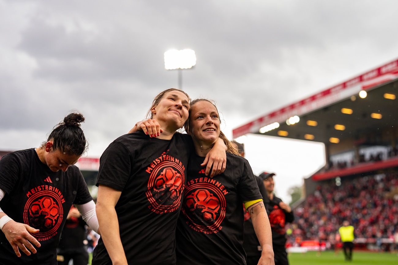 Zwei Fußballspielerinnen in schwarzen T-Shirts umarmen sich lächelnd, während sie das Stadion betreten. Wolken am Himmel.