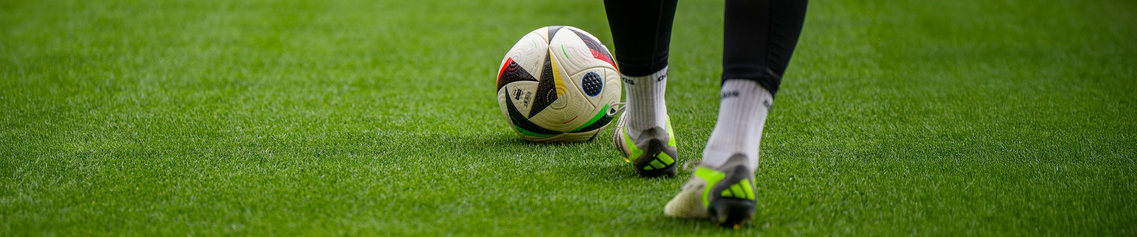 Ein Fußball rollt über einen grünen Rasen, während eine Spielerfigur mit schwarzen Hosen und weißen Socken in der Nähe steht.