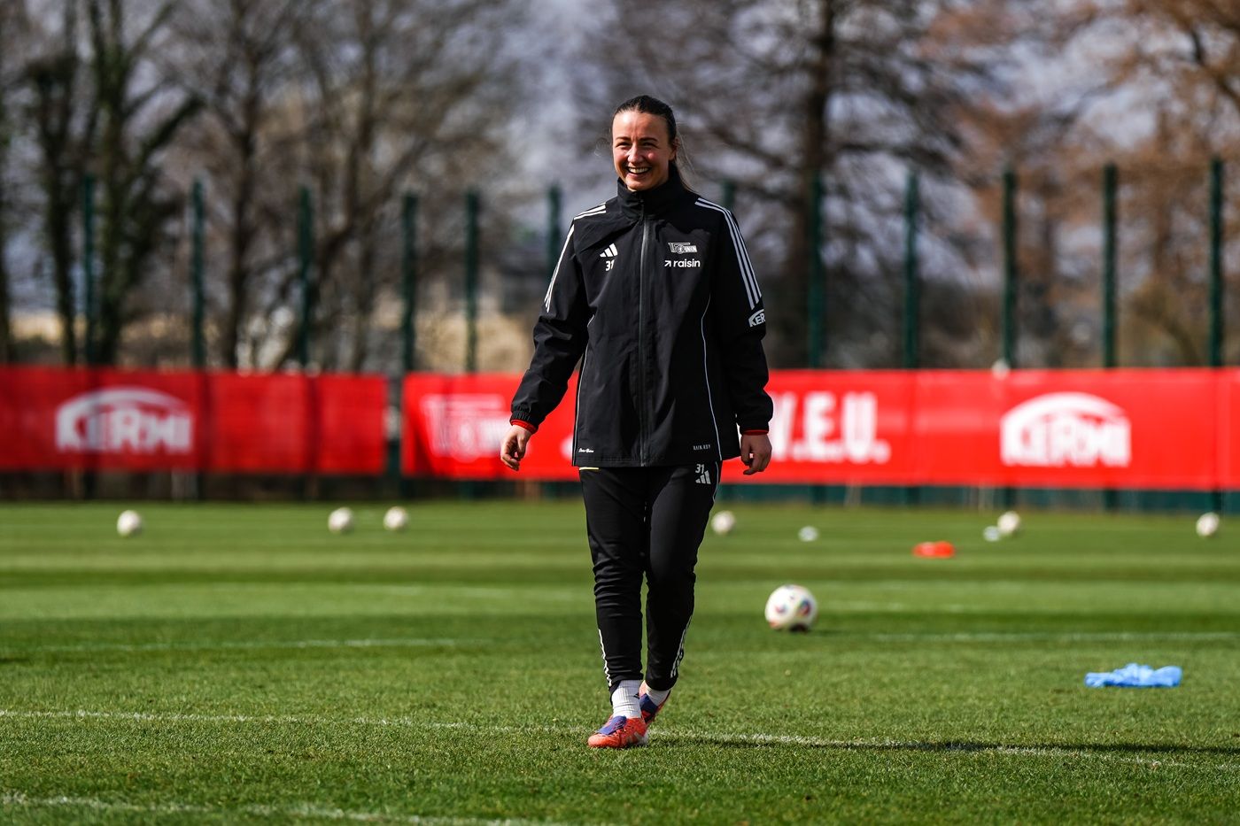 Eine Person in einem schwarzen Trainingsanzug lächelt und läuft über ein Fußballfeld, während Bälle im Hintergrund liegen.