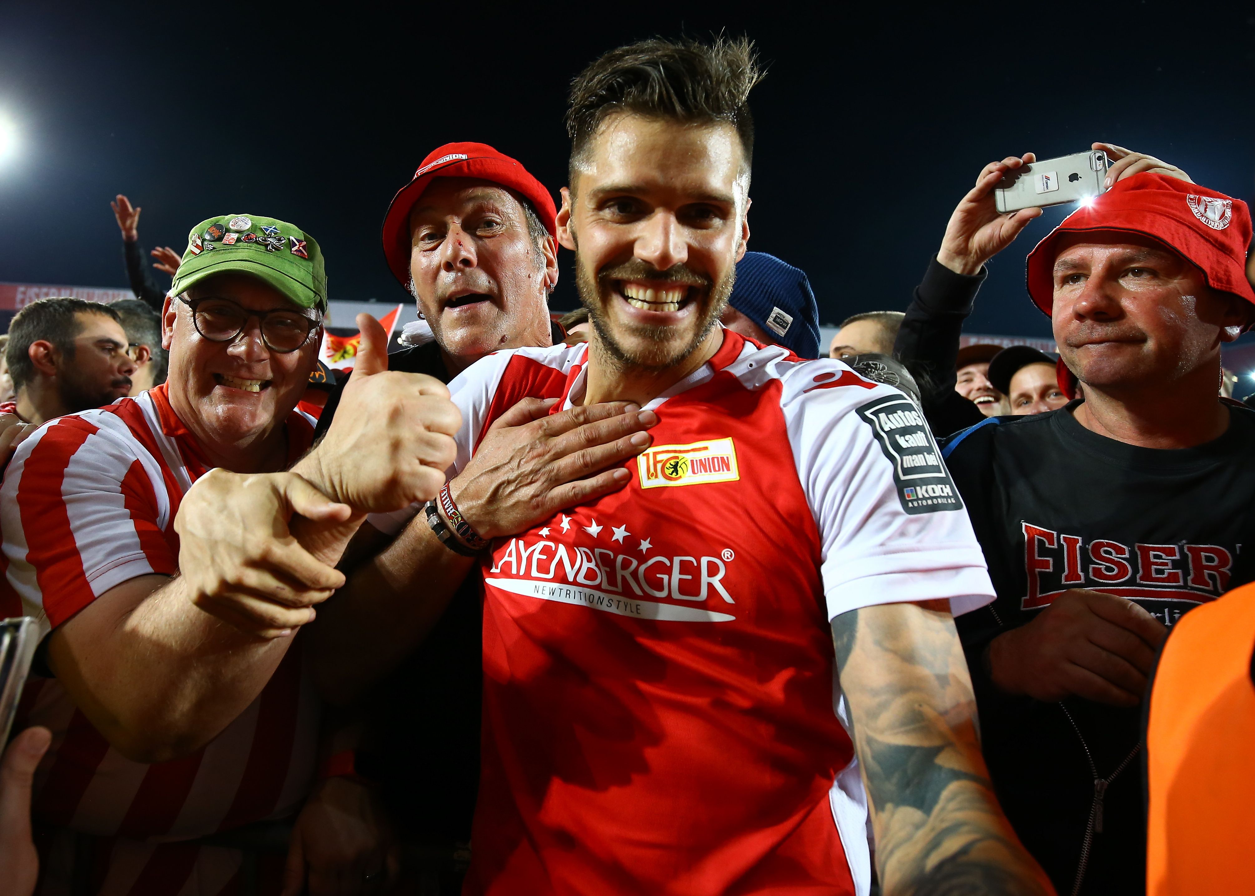 Ein Fußballspieler feiert mit jubelnden Fans in der Menge, während er lächelt und eine Hand auf seinem Herzen platziert.