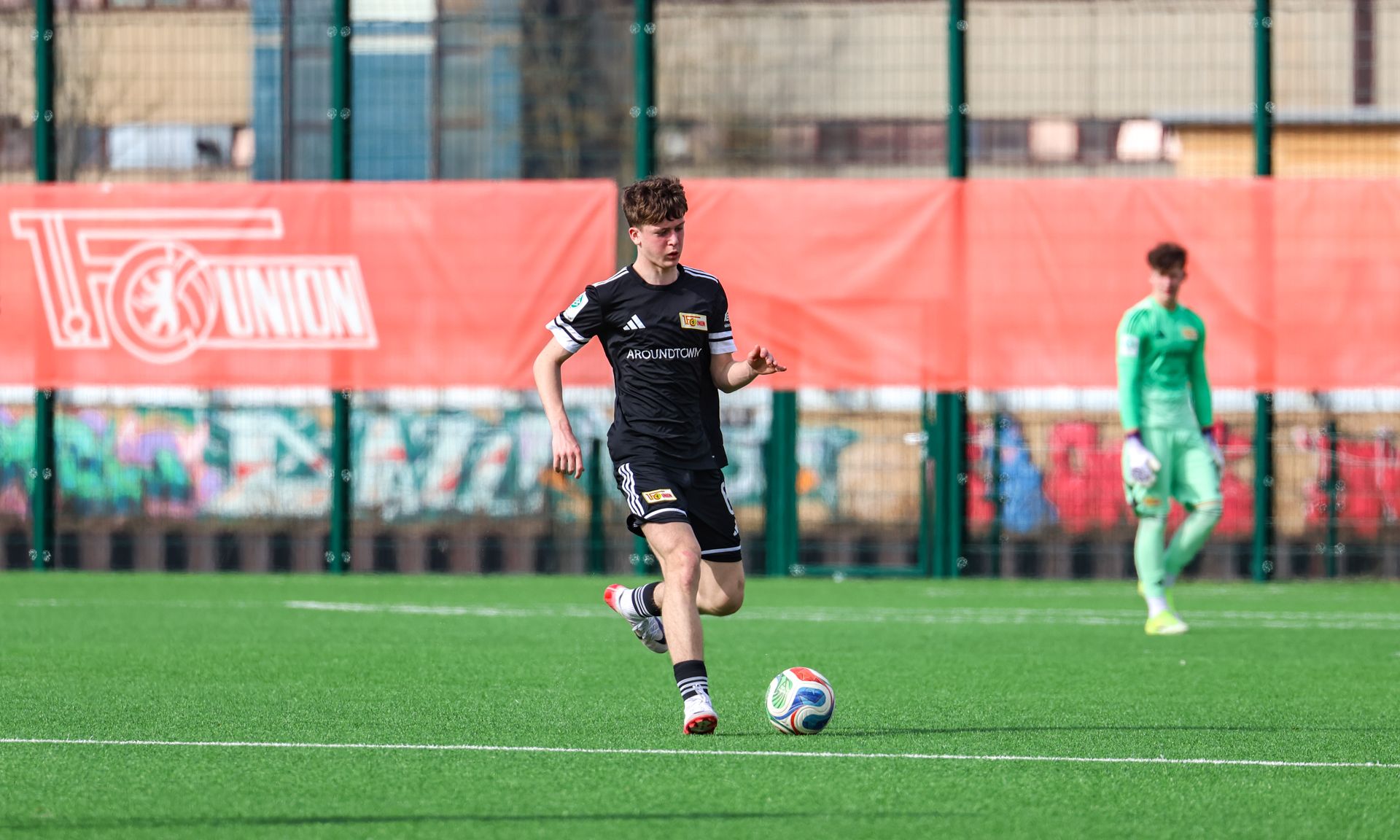 Ein junger Fußballspieler in schwarzer Shorts und Trikot läuft mit dem Ball auf einem Kunstrasenplatz. Im Hintergrund steht ein Torwart.