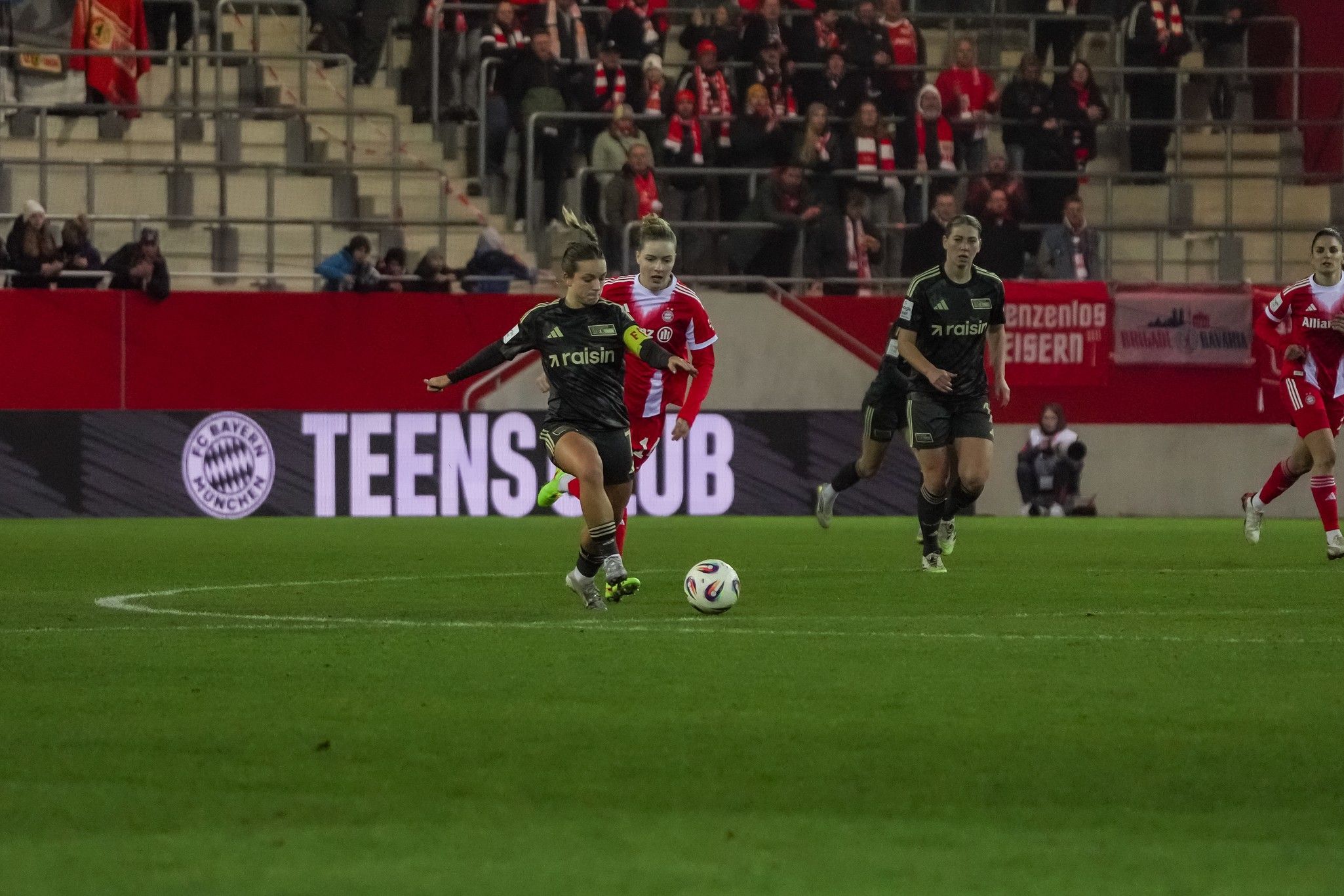 Eine Frau im Fußballtrikot spielt den Ball auf einem Stadionfeld, während Zuschauer im Hintergrund cheering.