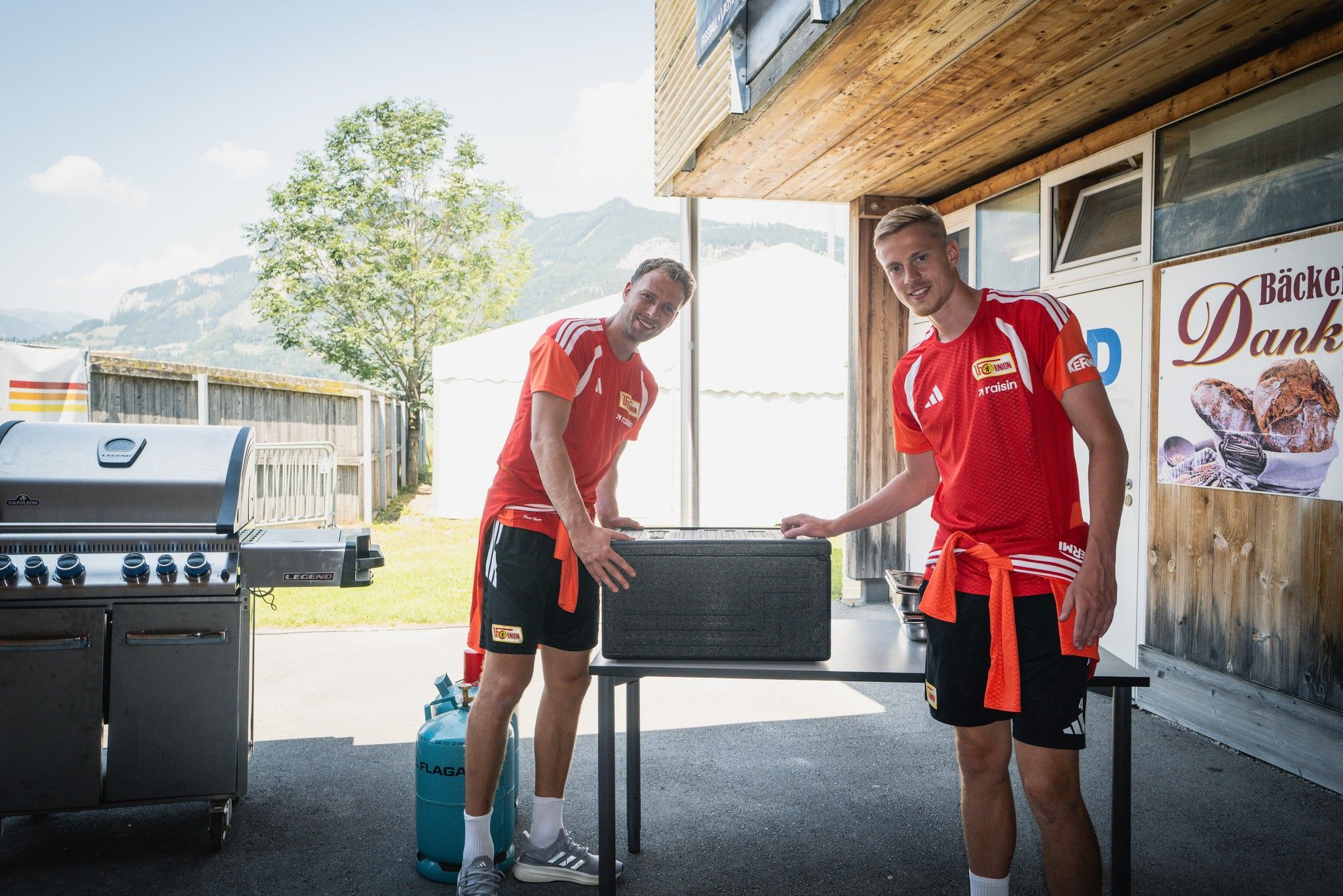 Zwei lächelnde Männer in roten Sporttrikots stehen neben einem Grill und einem Tisch im Freien. Im Hintergrund sind Berge zu sehen.