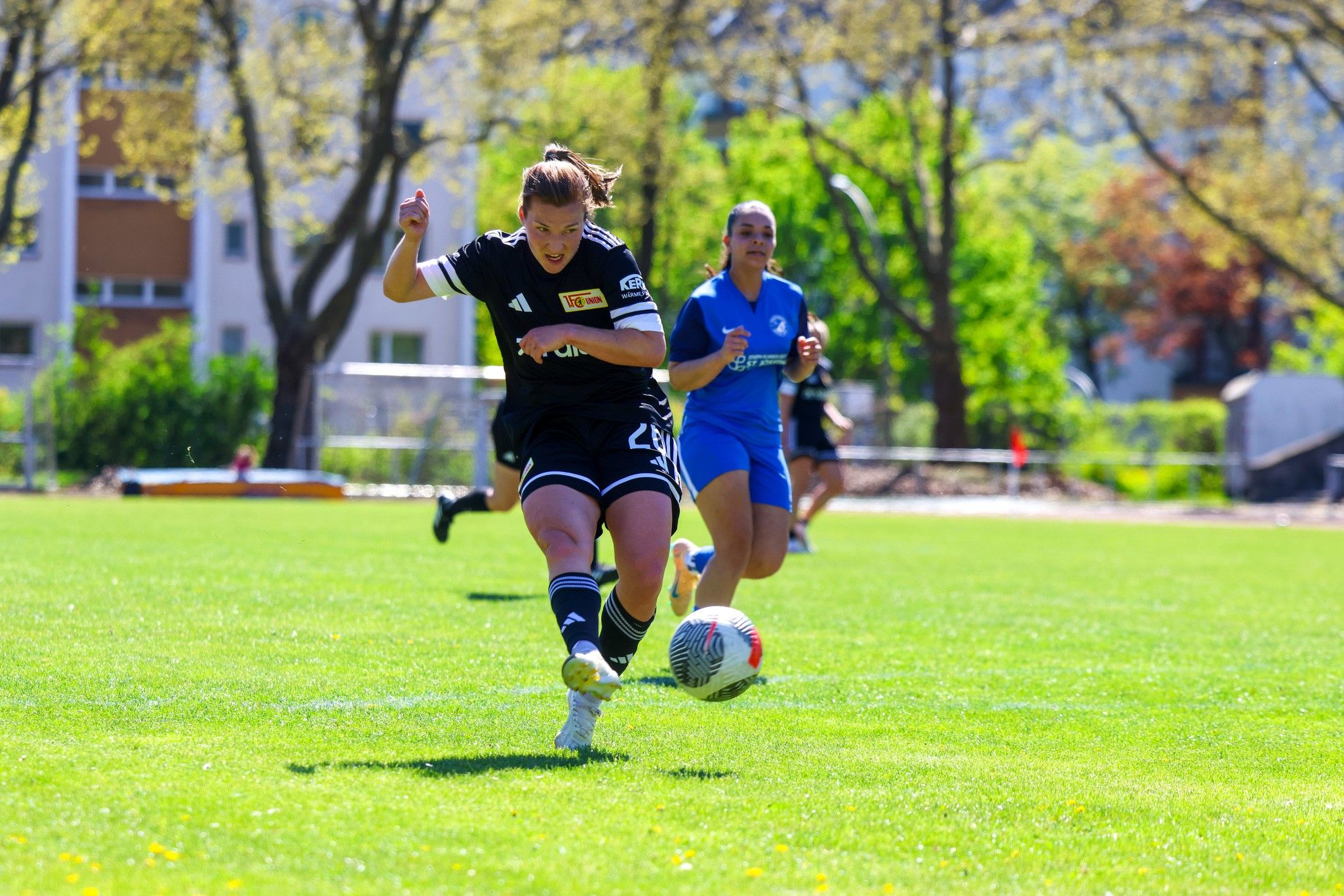 Zwei Frauen im Fußballtrikot spielen auf einem grünen Platz; eine Spielerin sprintet dem Ball entgegen.