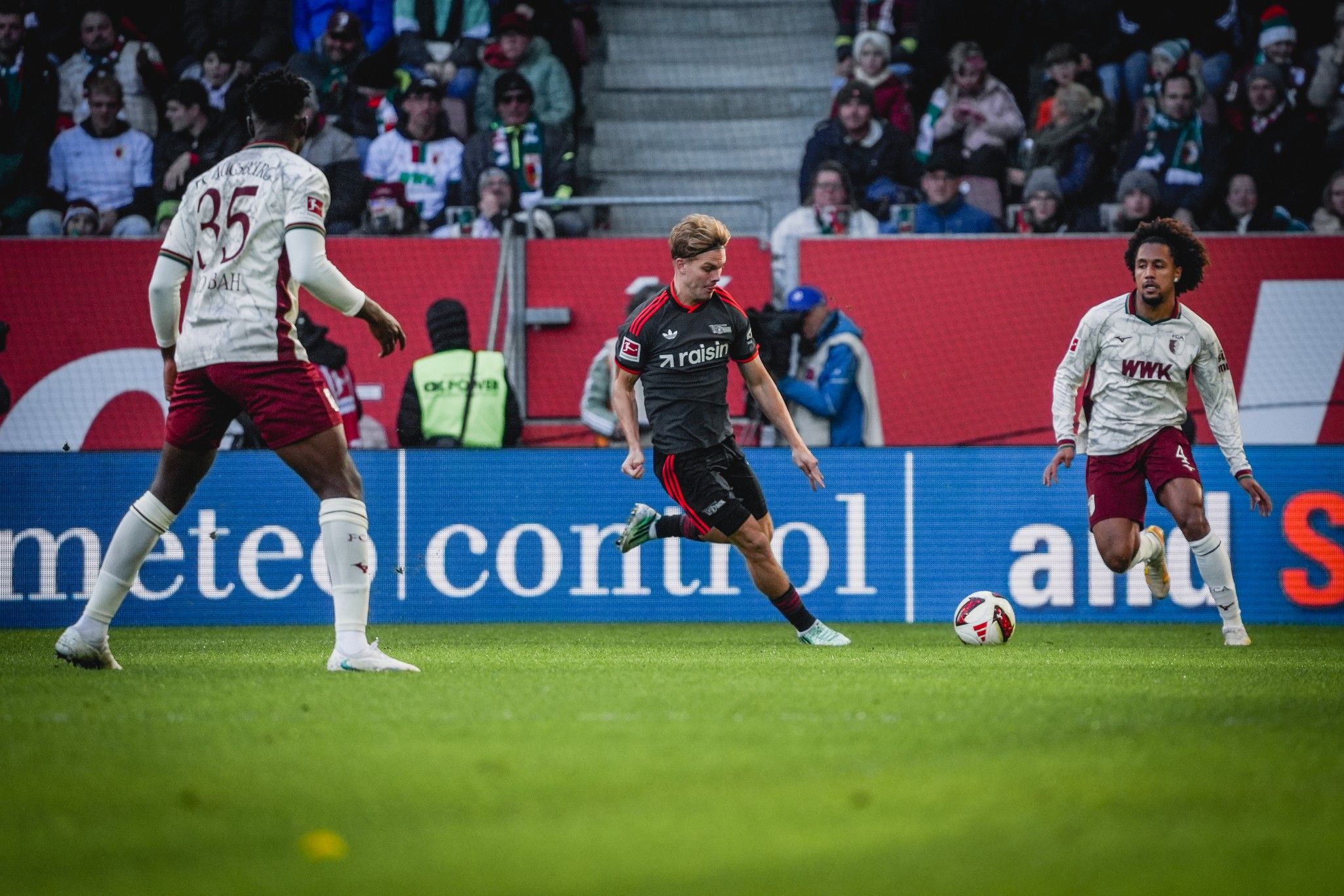 Ein Fußballspieler mit blondem Haar dribbelt den Ball, während zwei Gegenspieler in der Nähe stehen. Fans sind im Hintergrund zu sehen.