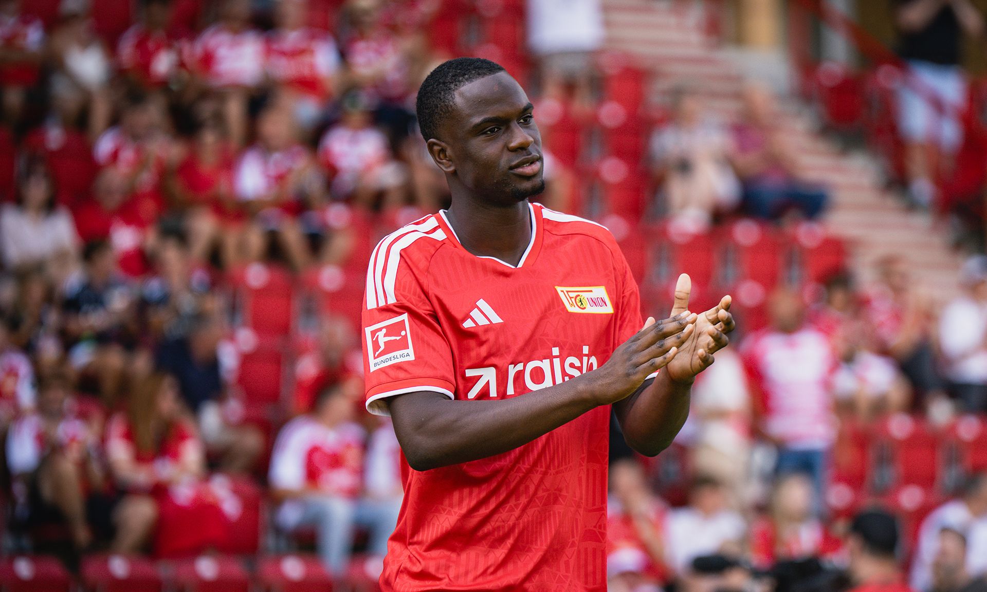 Ein Fußballspieler in rotem Trikot applaudiert in einem Stadion mit jubelnden Fans im Hintergrund.