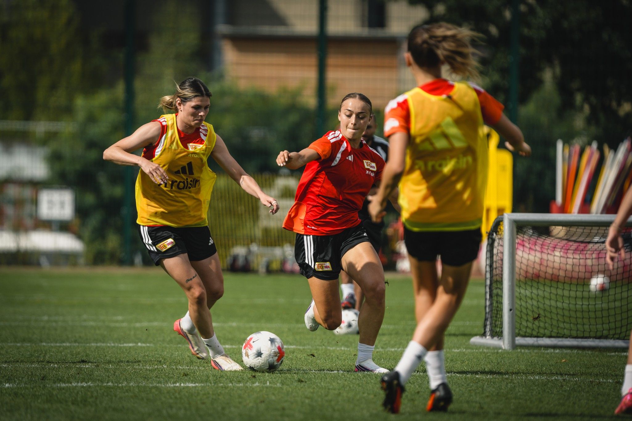 Zwei weibliche Fußballspielerinnen des Teams trainieren auf dem Rasen, während eine dritte Spielerin den Ball dribbelt.
