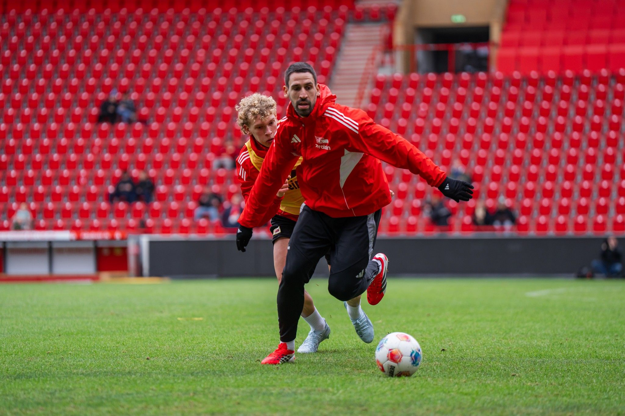 Zwei Fußballspieler im Training, einer dribbelt den Ball, der andere versucht, ihn zu erreichen. Leere Tribüne im Hintergrund.
