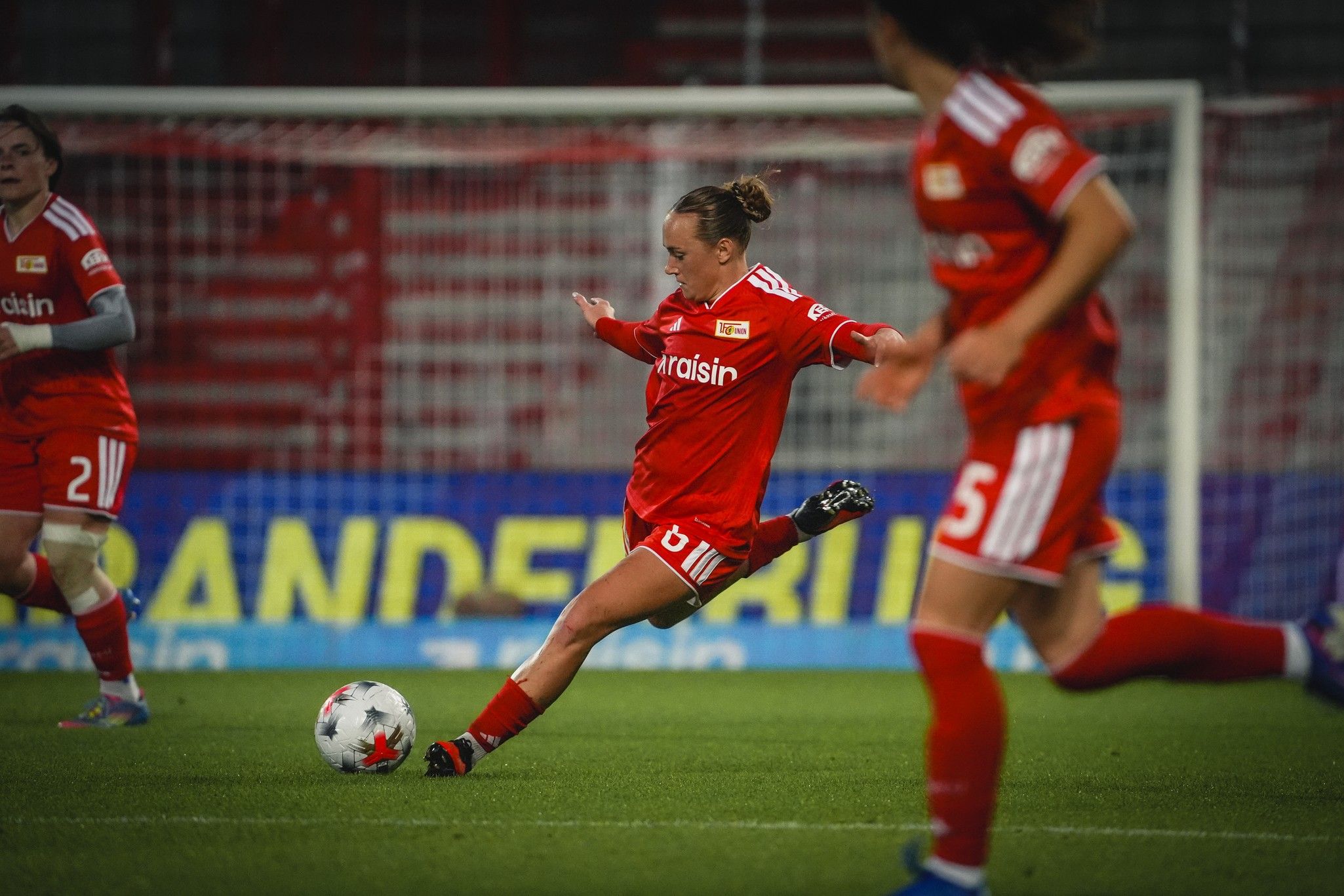 Eine Spielerin in rotem Trikot schießt den Ball im Fußballspiel, während zwei Teamkolleginnen im Hintergrund stehen.