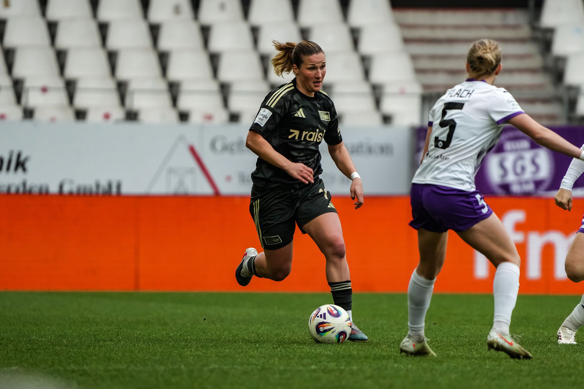 Eine Fußballspielerin in schwarzem Trikot dribbelt den Ball, während eine Gegnerin in weißem Trikot interessiert zusieht.