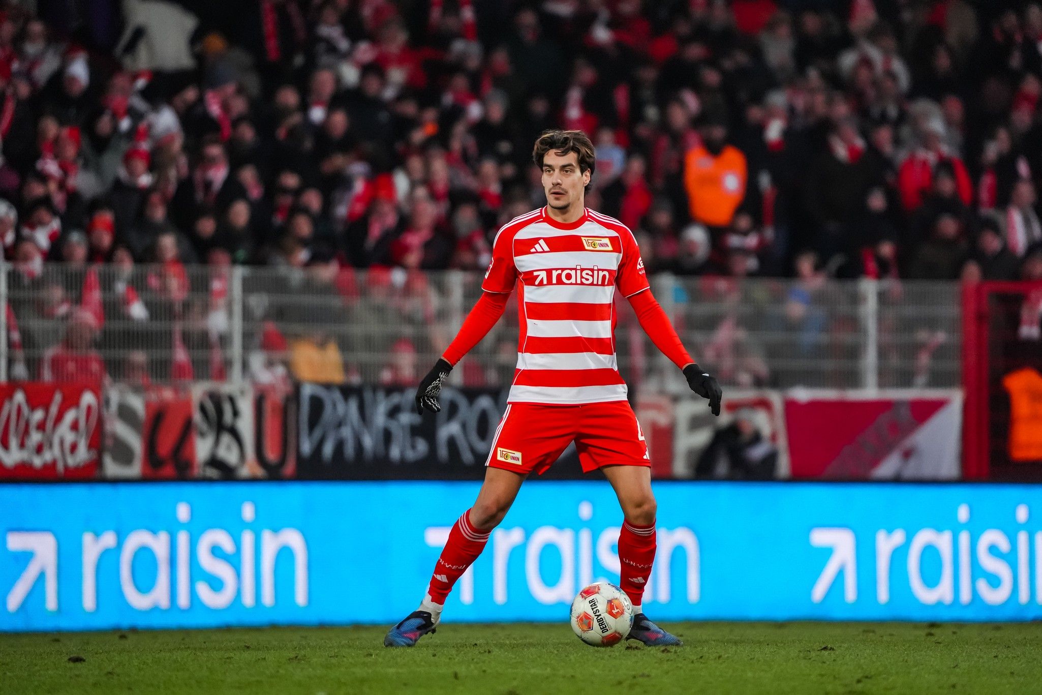 Ein Fußballspieler in rot-gestreifter Uniform steht auf dem Spielfeld, umgeben von jubelnden Fans im Hintergrund.