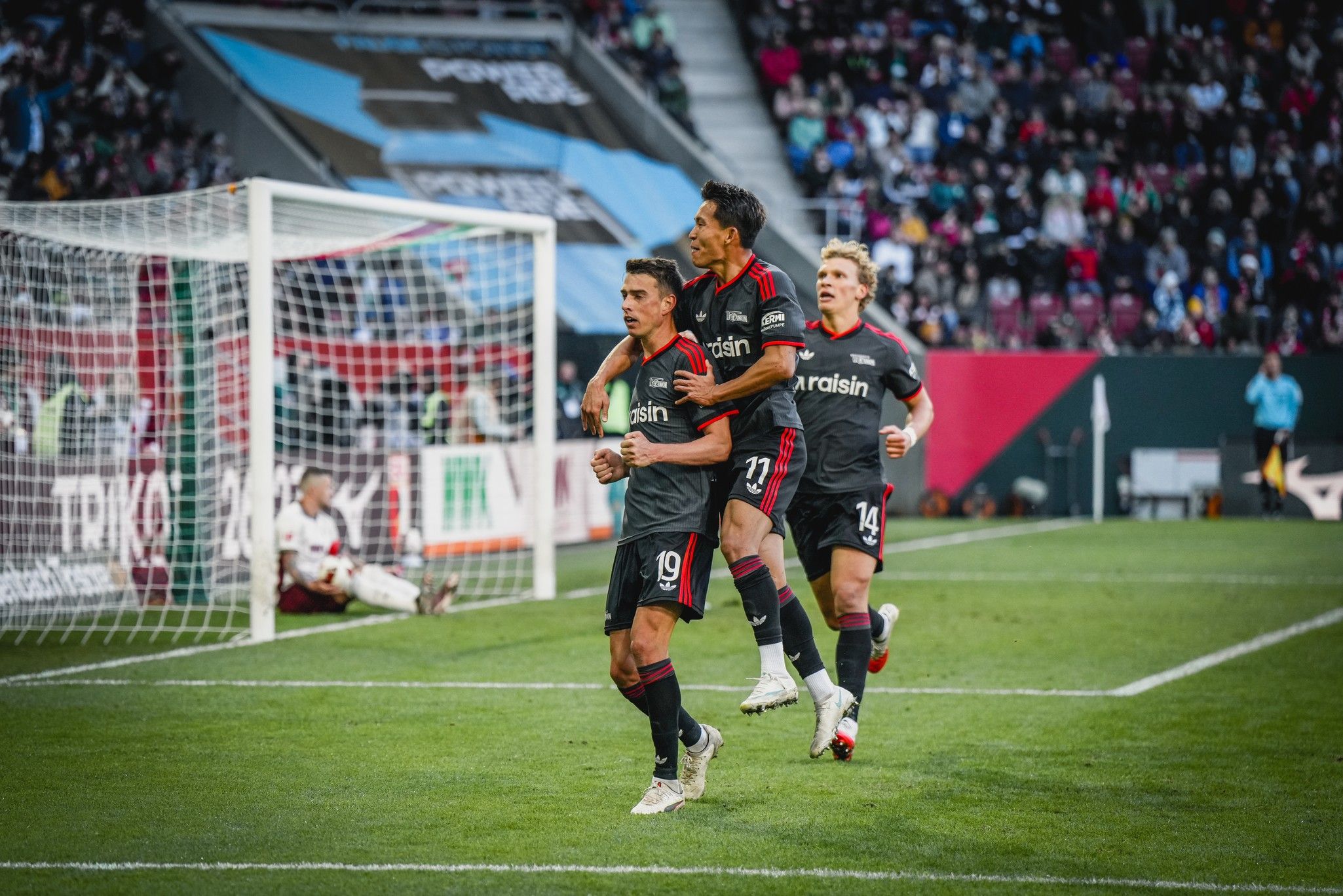 Drei Fußballspieler jubeln nach einem Tor, während ein Gegner enttäuscht im Hintergrund sitzt.